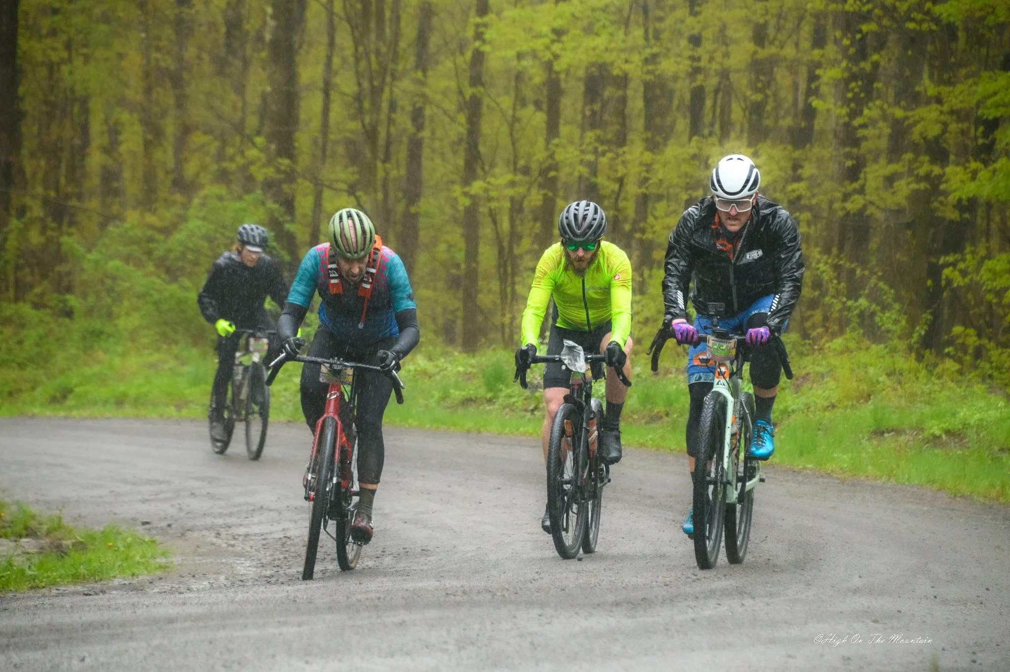 Four cyclists riding on a dirt trail through a green, wooded area, wearing helmets and rain gear.