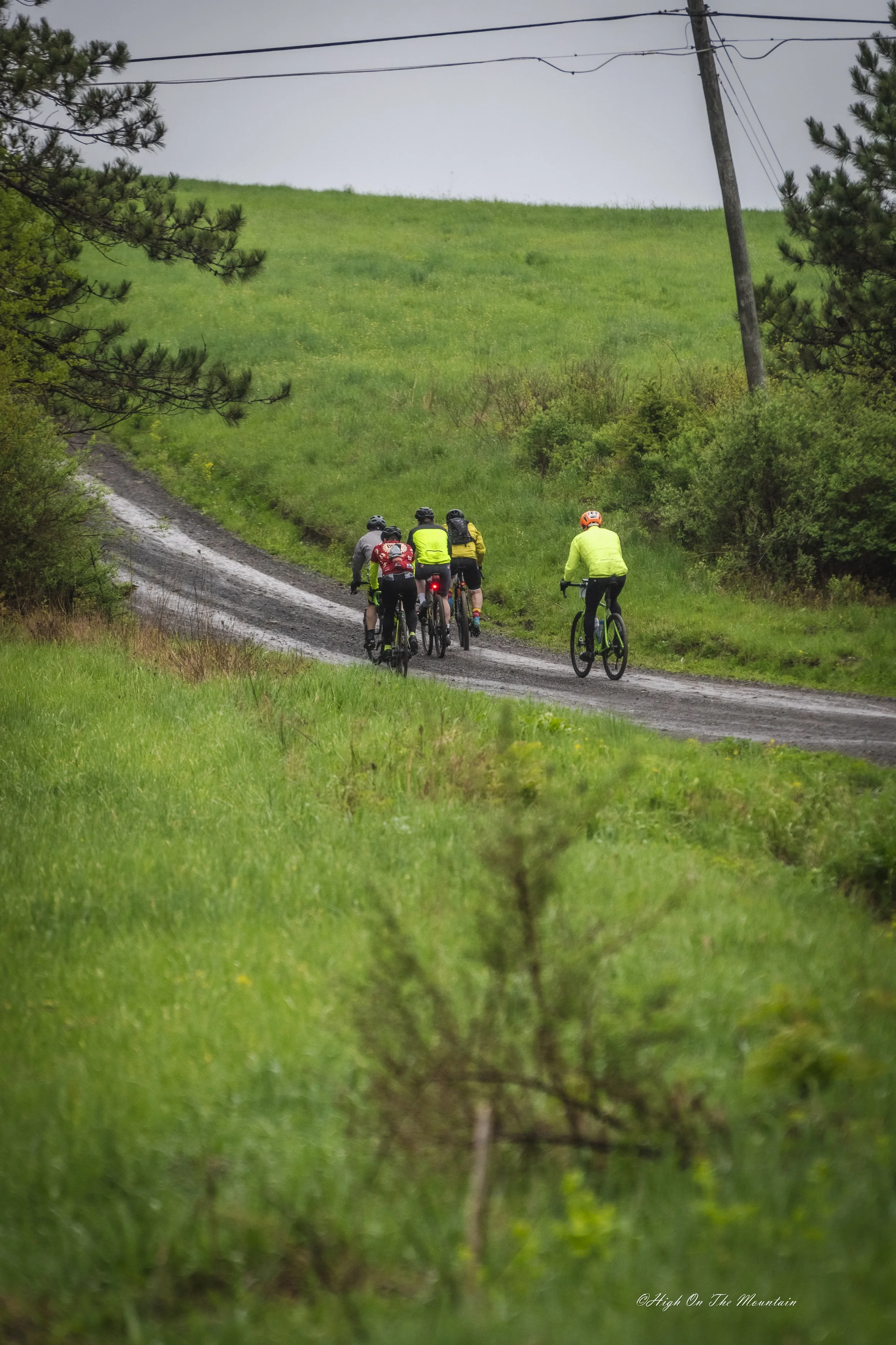 A group of five cyclists wearing helmets and bright clothing riding on a muddy dirt trail, surrounded by green grass and bushes on a cloudy day.