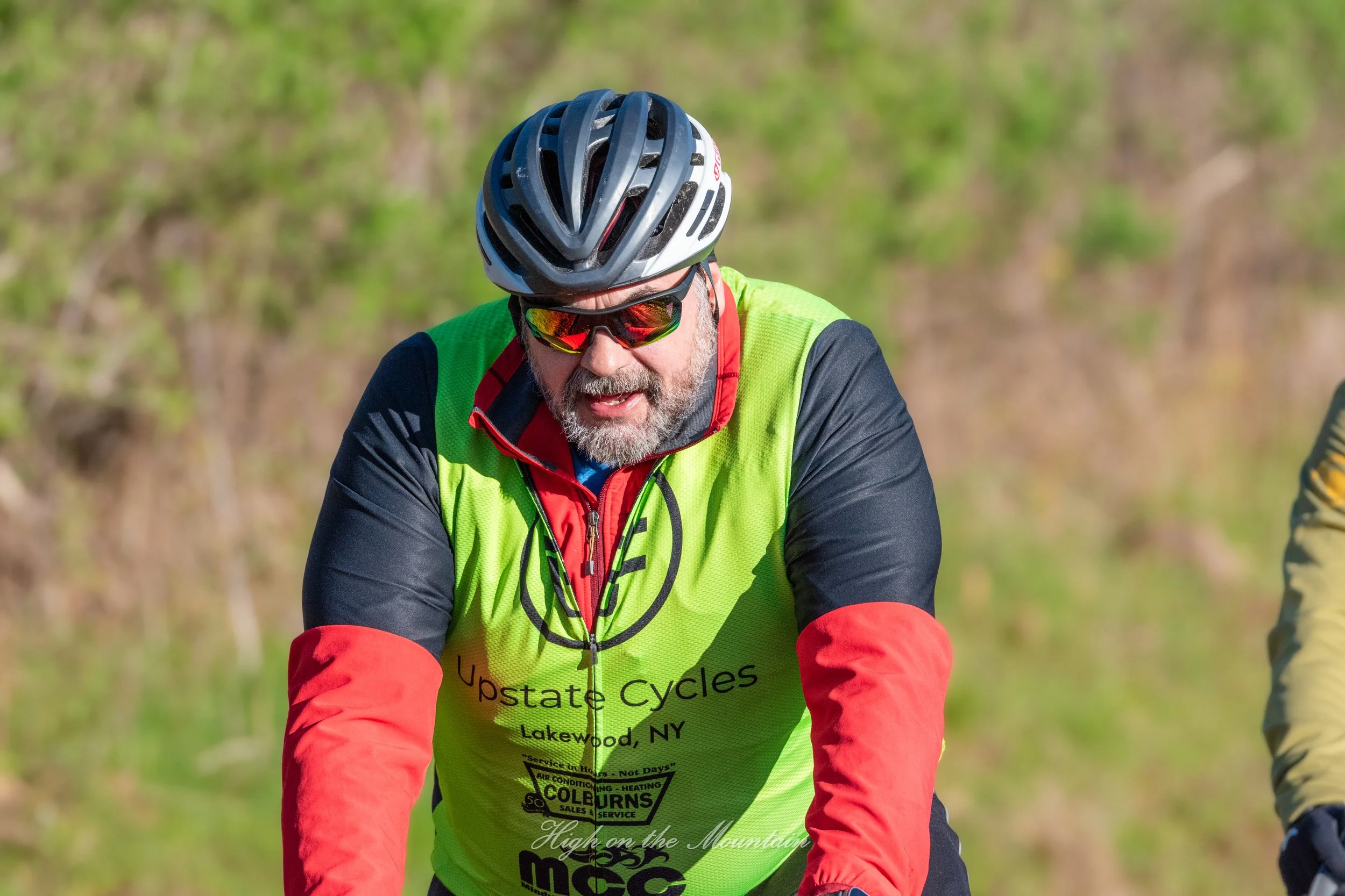 A man riding a bicycle outdoors on a sunny day, wearing a black helmet, colorful reflective sunglasses, a bright green cycling vest with advertisements, and a red and black long sleeve shirt.