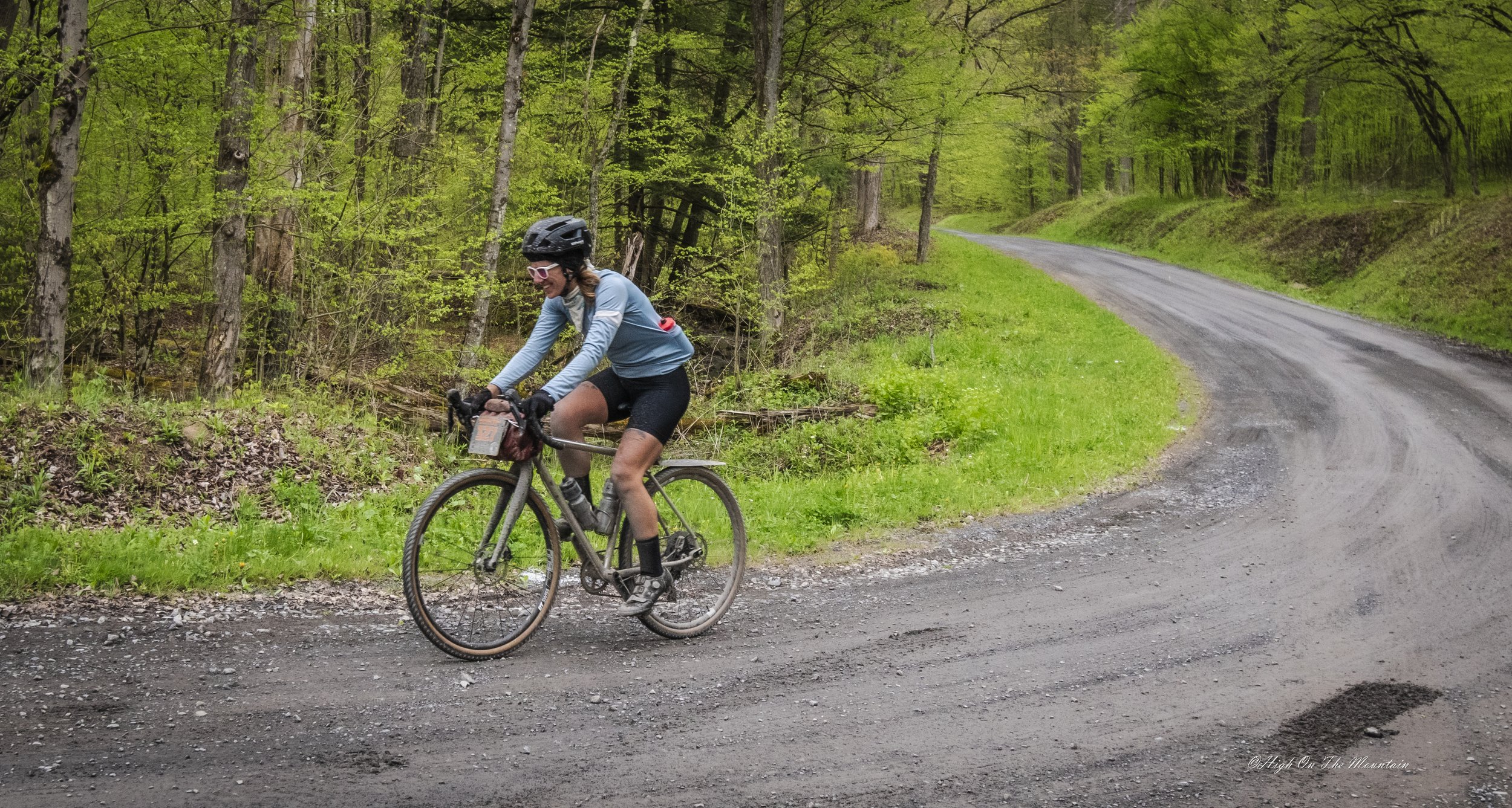 A woman riding a mountain bike along a dirt trail in a lush green forest, wearing a helmet, sunglasses, and a light blue jacket.