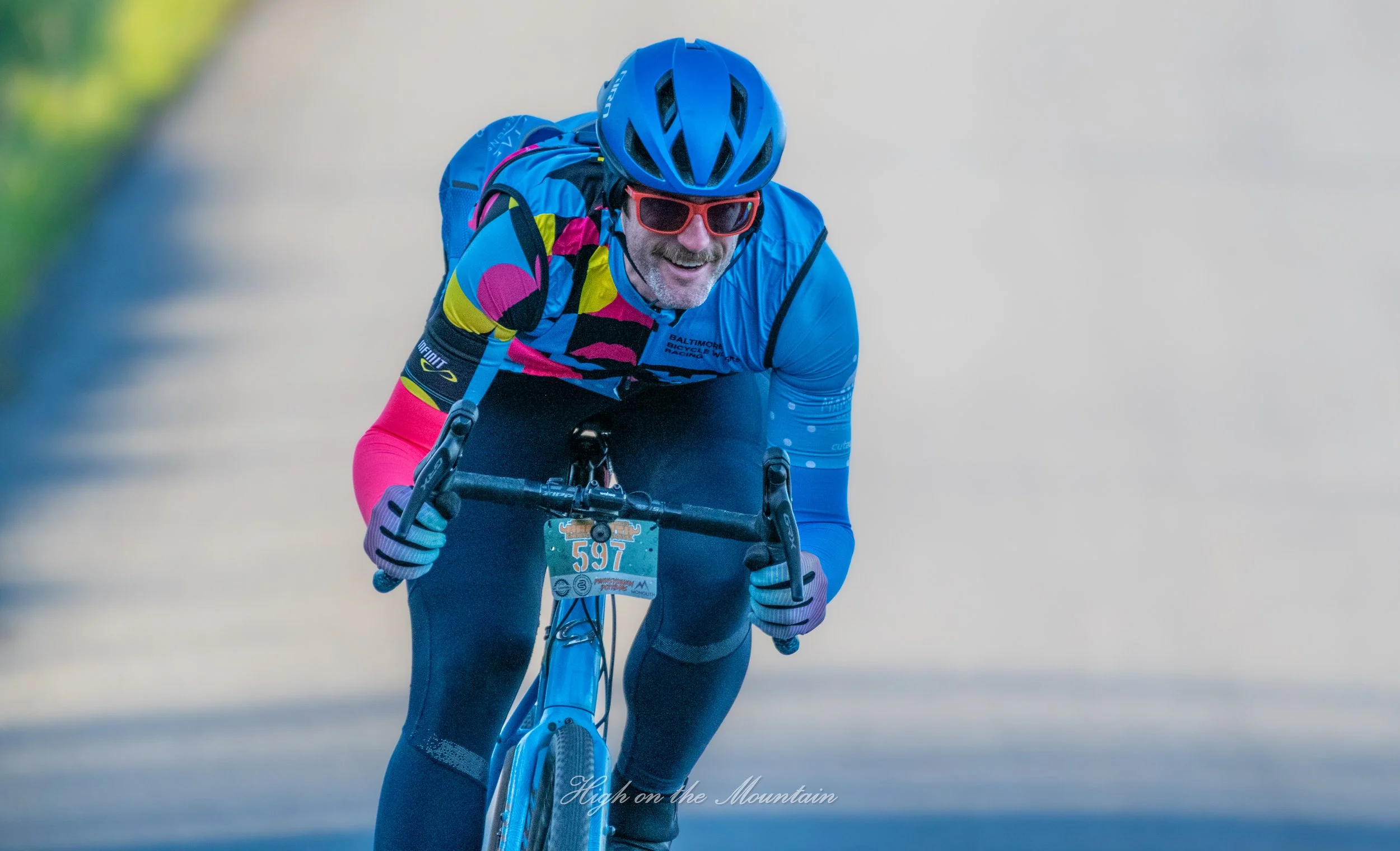A male cyclist wearing a blue helmet, colorful cycling jersey, sunglasses, and gloves, riding a mountain bike on a trail. He is smiling and appears to be enjoying the ride.