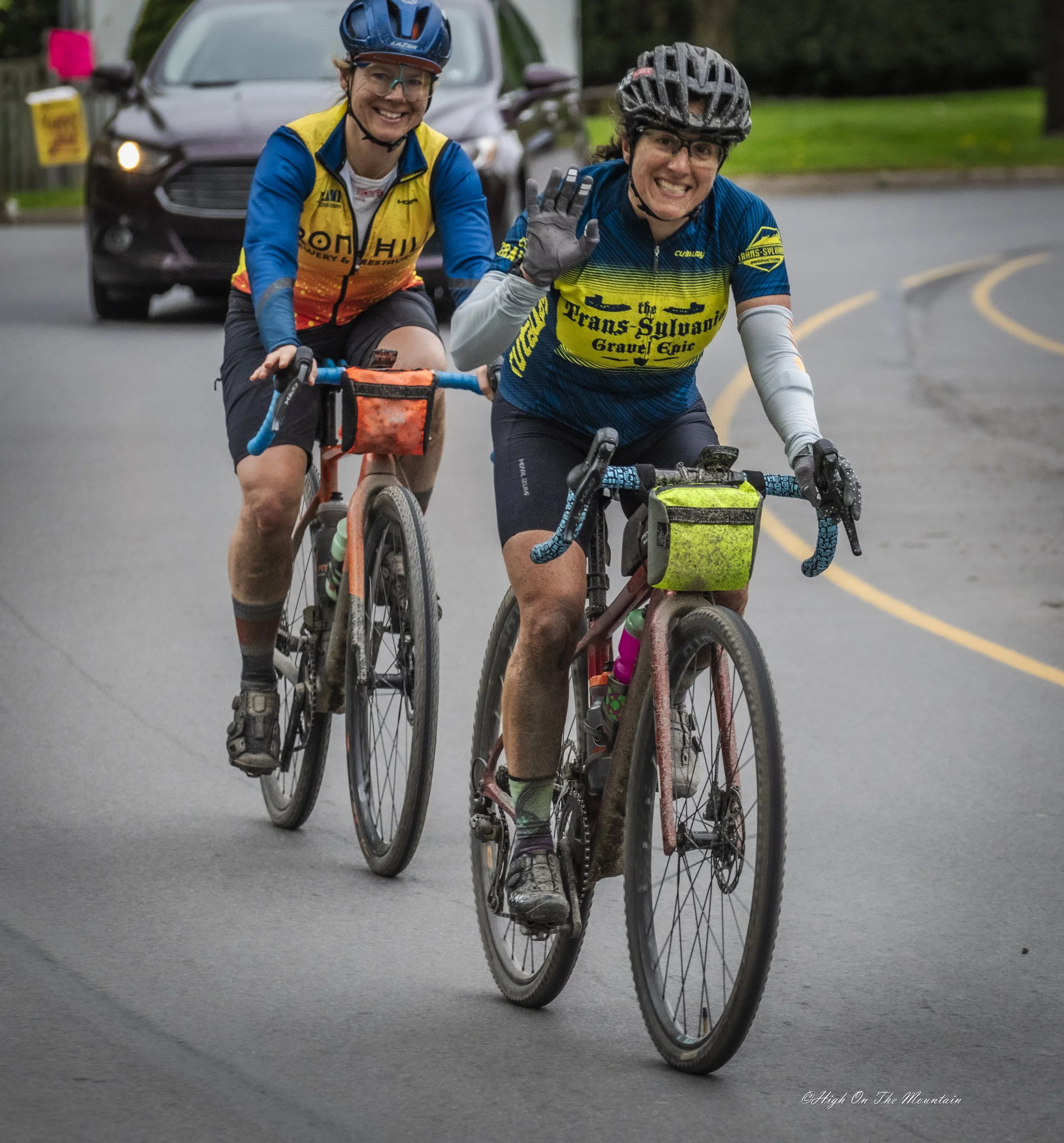 Two women riding bicycles on a paved road, both wearing helmets and cycling gear, smiling and waving at the camera, with a car and greenery in the background.