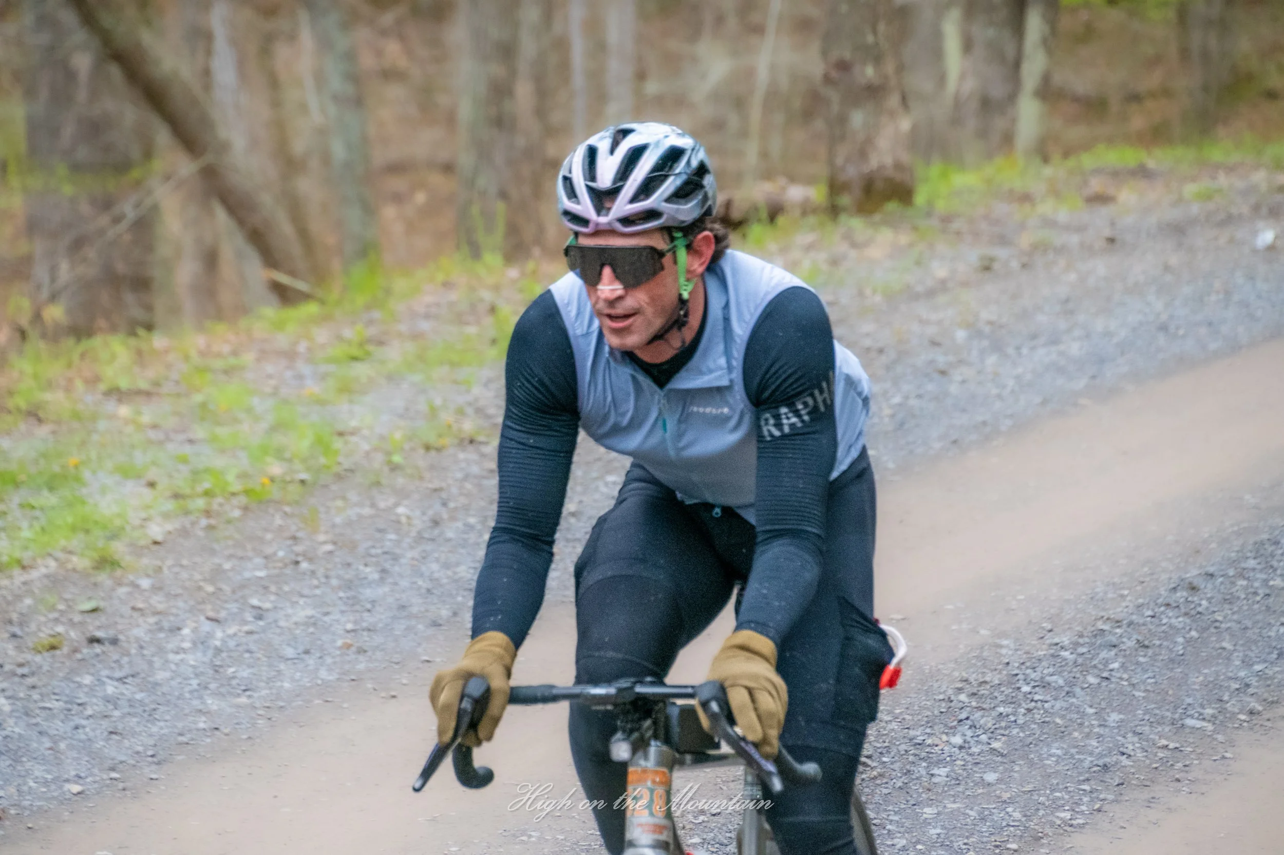 A person riding a mountain bike on a dirt trail through a wooded area, wearing a helmet, sunglasses, gloves, and athletic clothing.