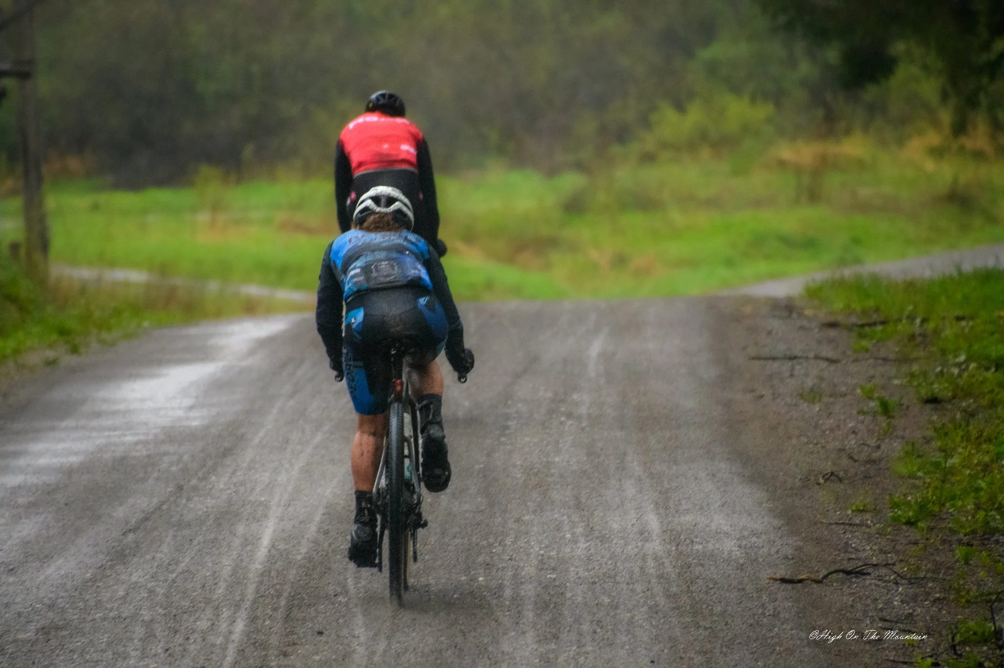 A cyclist riding on a gravel path in a green, wooded area during rainy weather, wearing a red and black jacket and helmet.