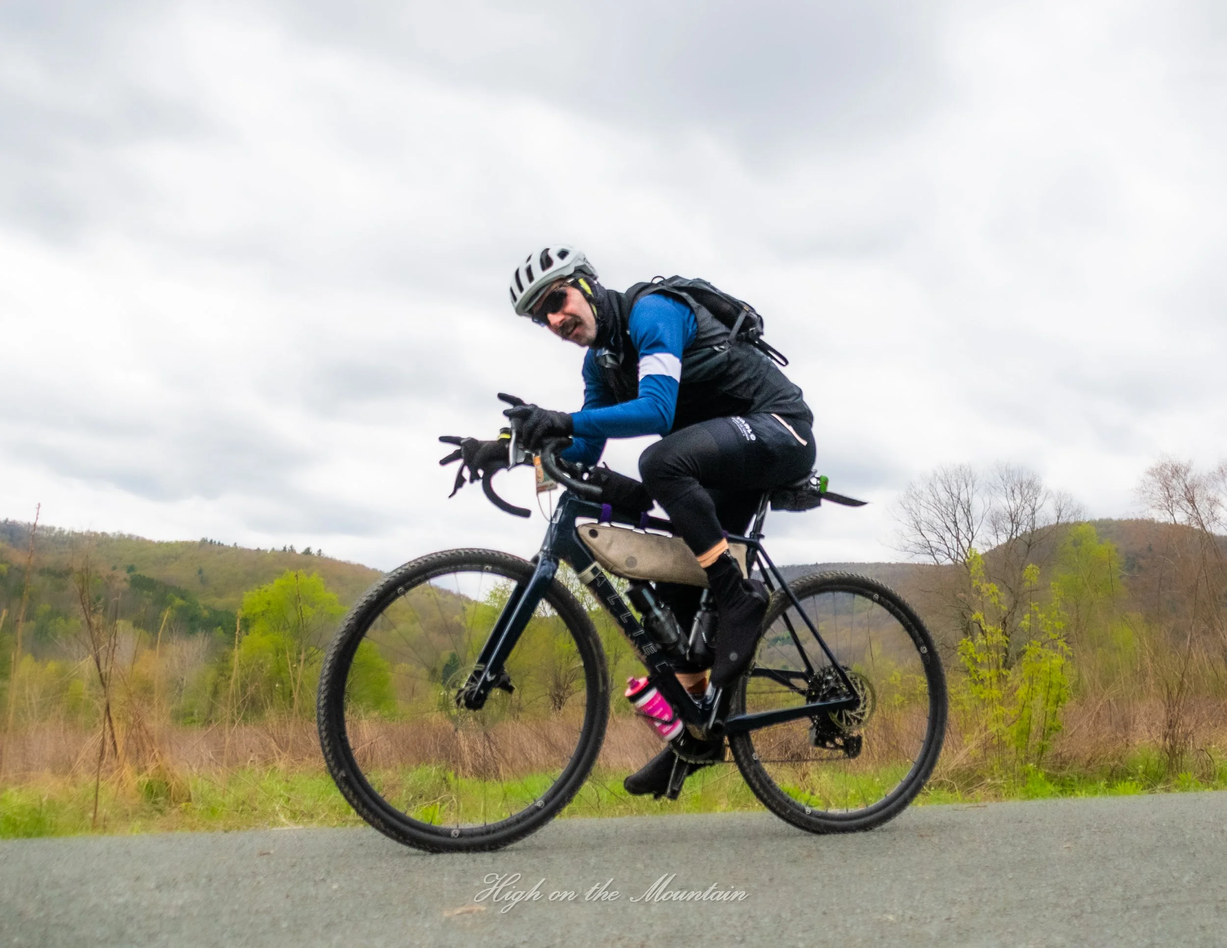 A man wearing a helmet and sunglasses riding a mountain bike on a trail with hills and trees in the background.