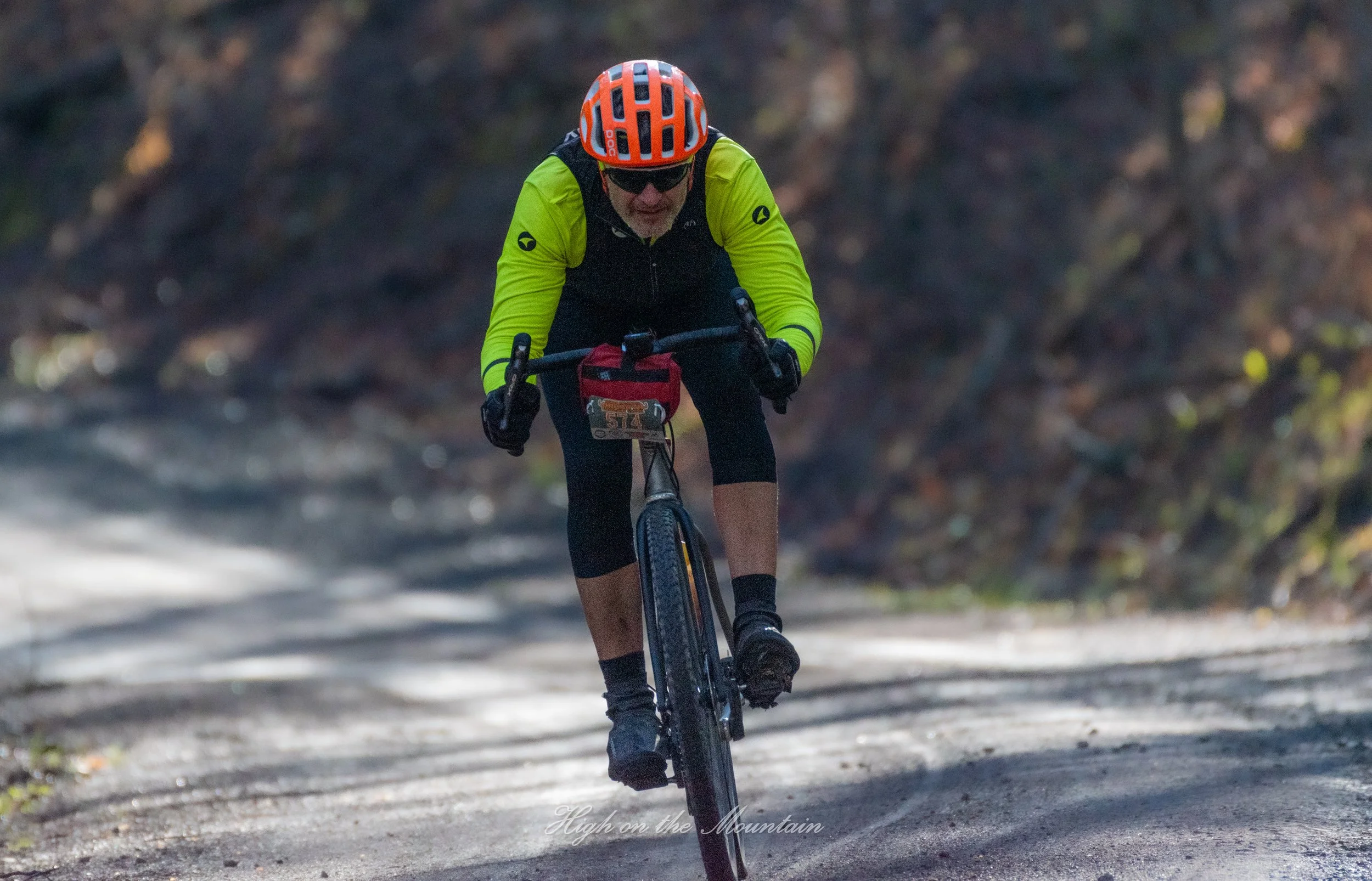 A person riding a mountain bike on a dirt trail through wooded area, wearing a bright yellow jacket, black helmet with orange accents, sunglasses, and black cycling gear.