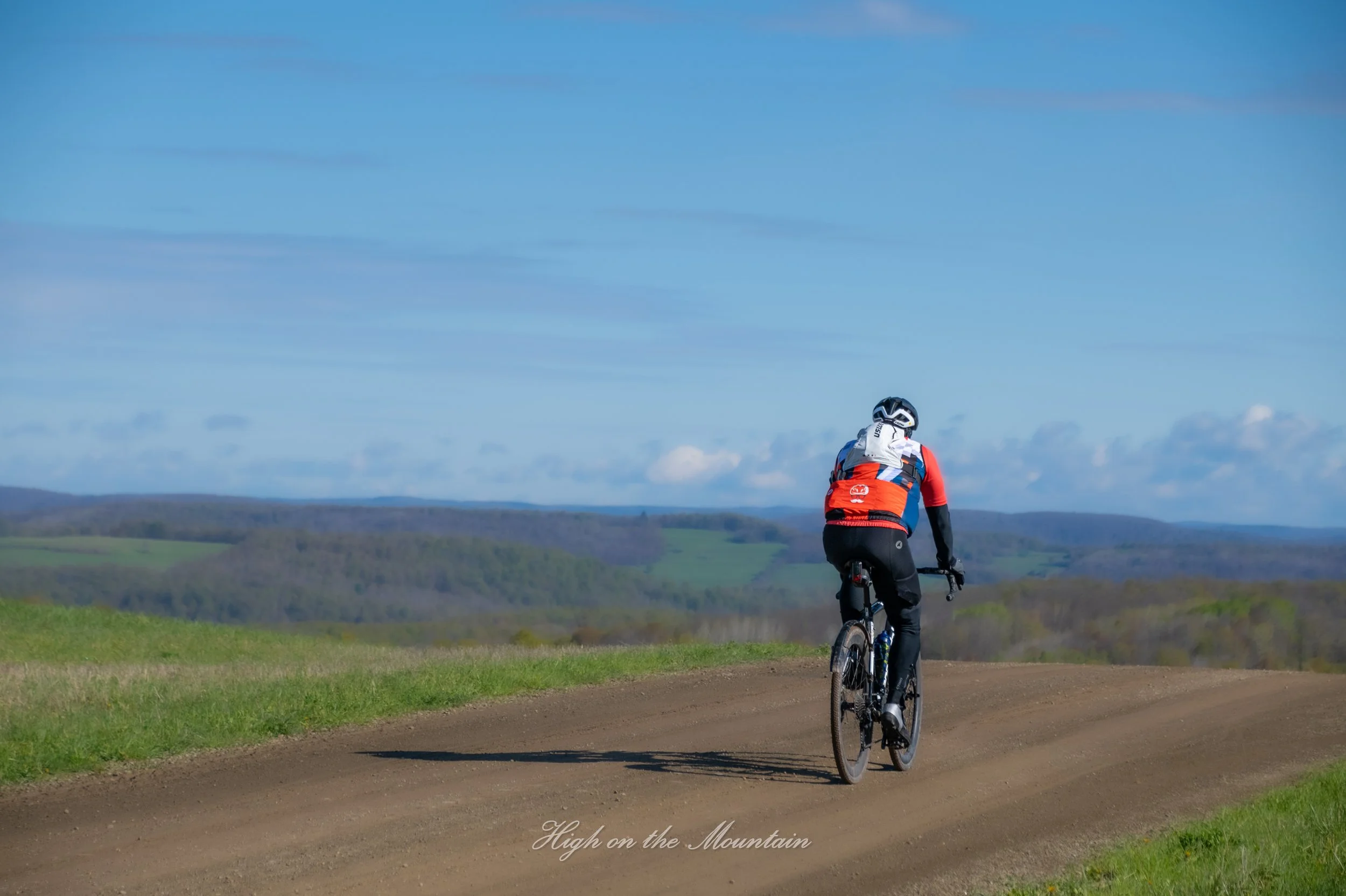 A person riding a mountain bike on a dirt path in a scenic rural landscape with rolling hills and a clear blue sky.