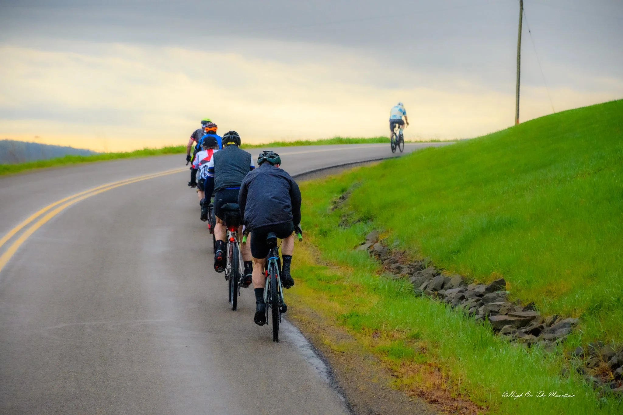 A group of cyclists riding uphill on a paved road with green grass on the side, sky with clouds in the background, and a mountain cyclist at the top of the hill.