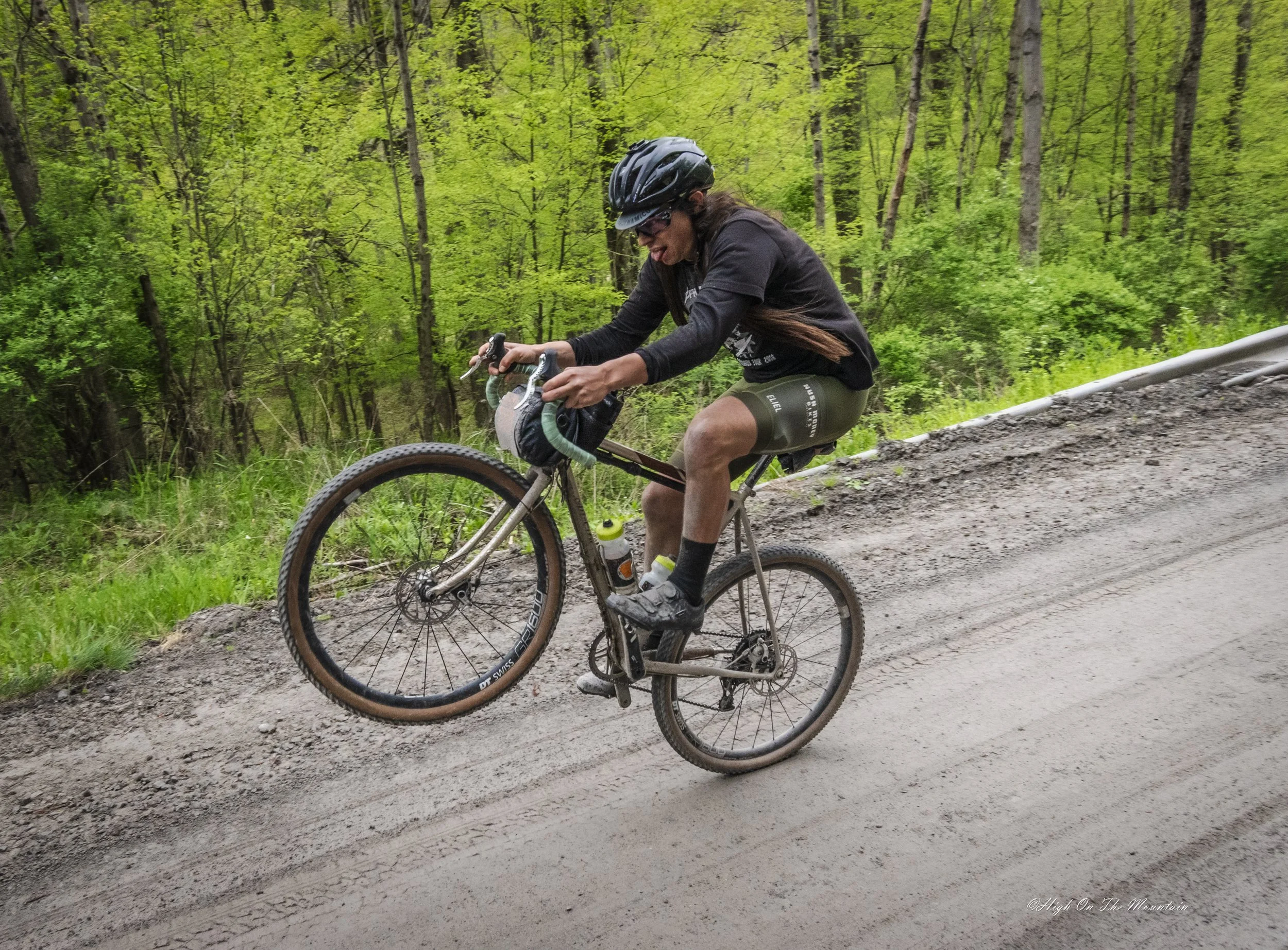 Person riding mountain bike on a dirt trail in a lush green forest wearing a black helmet, sunglasses, a black long-sleeve shirt, and olive green shorts.
