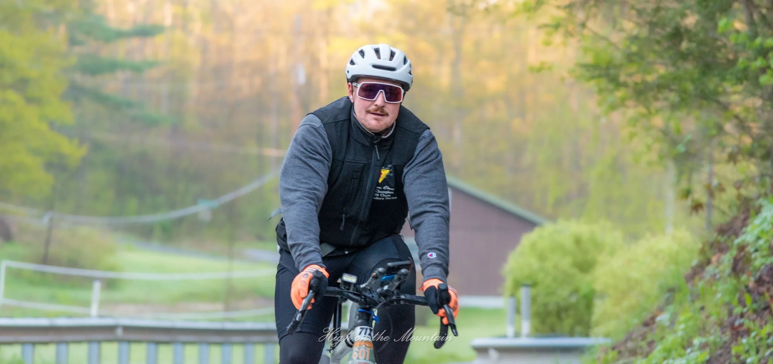 A man wearing a bike helmet and sunglasses riding a mountain bike in a green outdoor setting with trees and a house in the background.