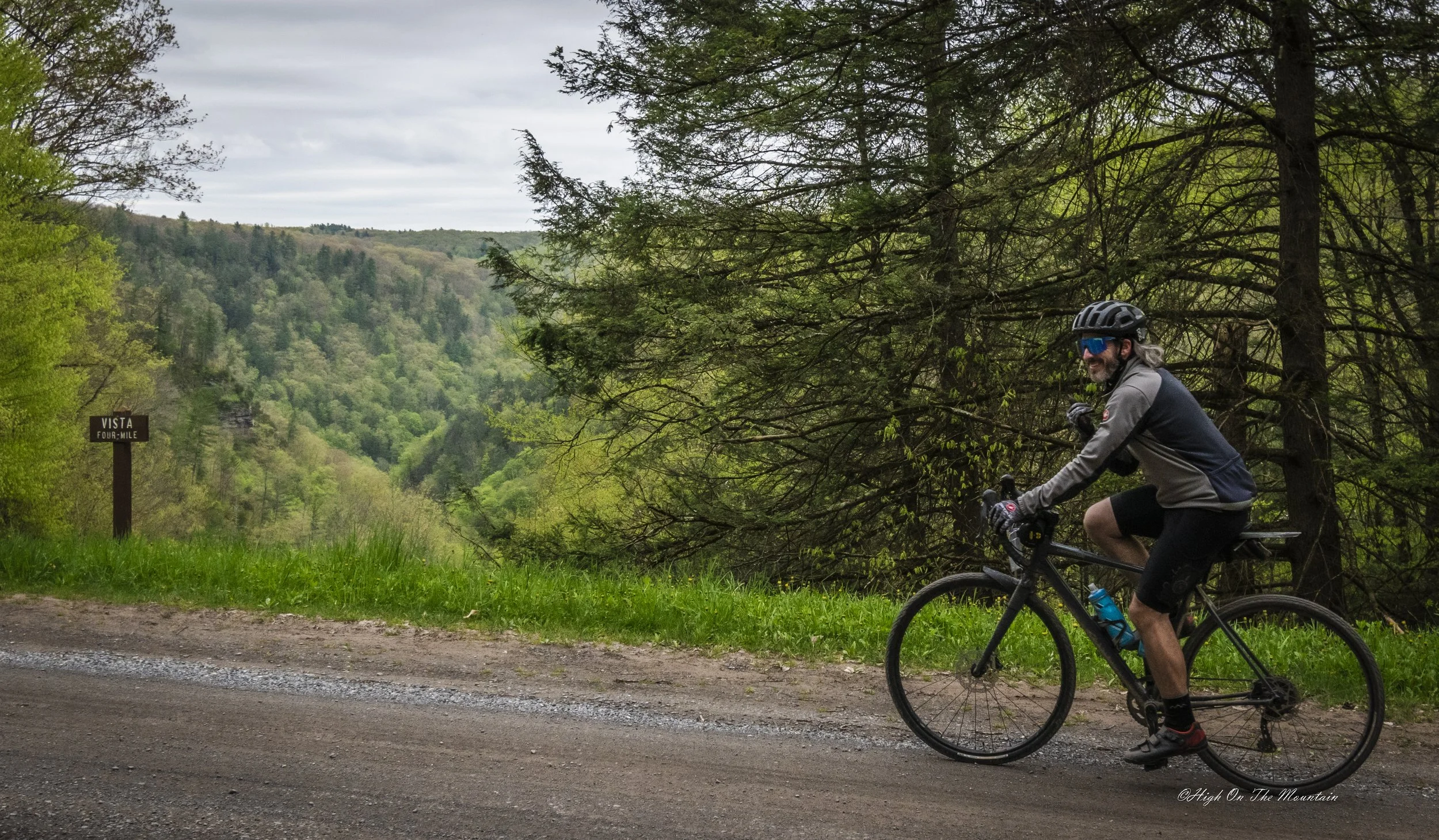A man riding a mountain bike on a trail by a lush green forest, with a sign indicating a four-mile vista. The man is wearing a helmet, sunglasses, a long sleeve jacket, and shorts, smiling while enjoying the outdoor ride.
