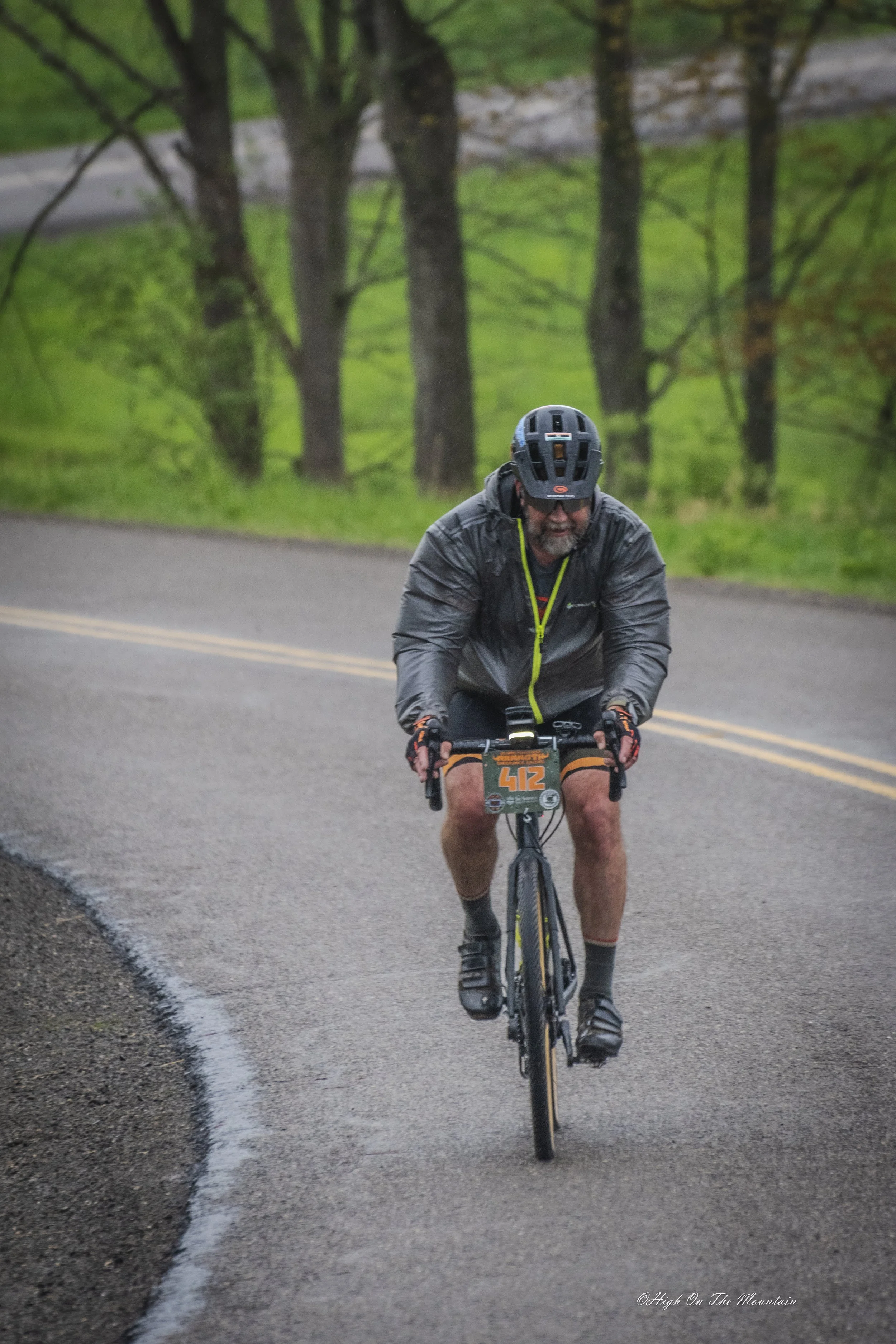 Man biking up a wet road on a rainy day, wearing a helmet and rain jacket, with trees and green landscape in the background.