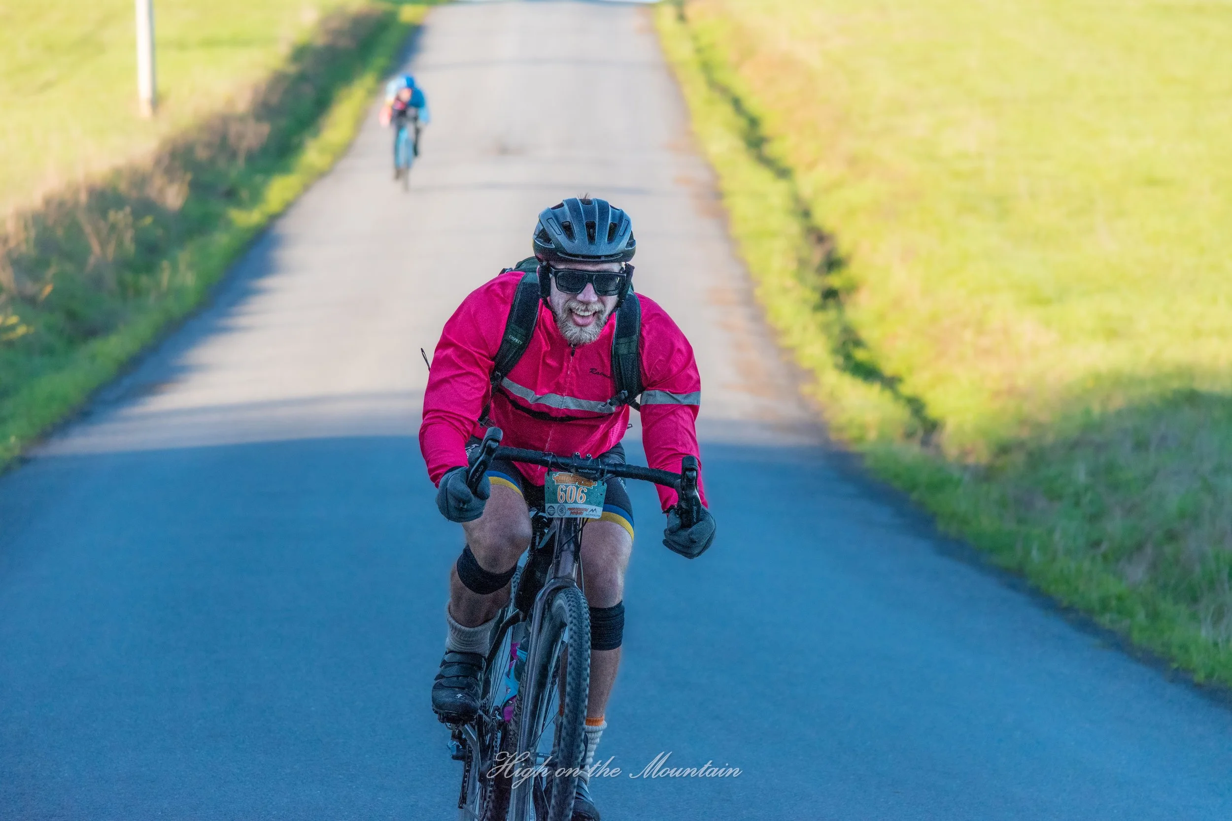 A man with a beard and sunglasses riding a mountain bike on a paved trail, wearing a black helmet and pink jacket, with another cyclist in the background on the same trail surrounded by green grass and trees.