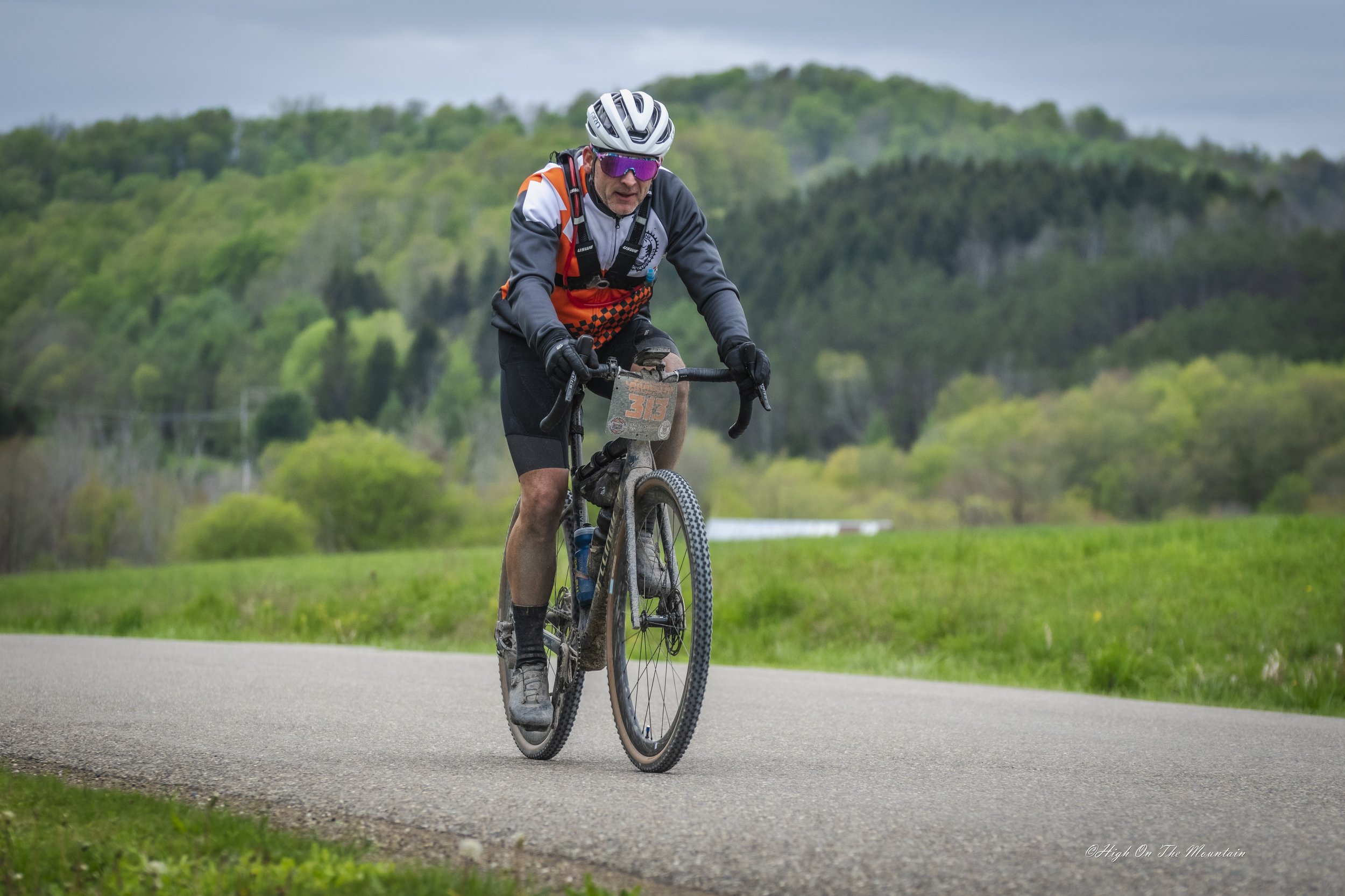 A man wearing a helmet, sunglasses, and cycling gear riding a mountain bike on a paved trail in a green landscape with hills and cloudy sky.