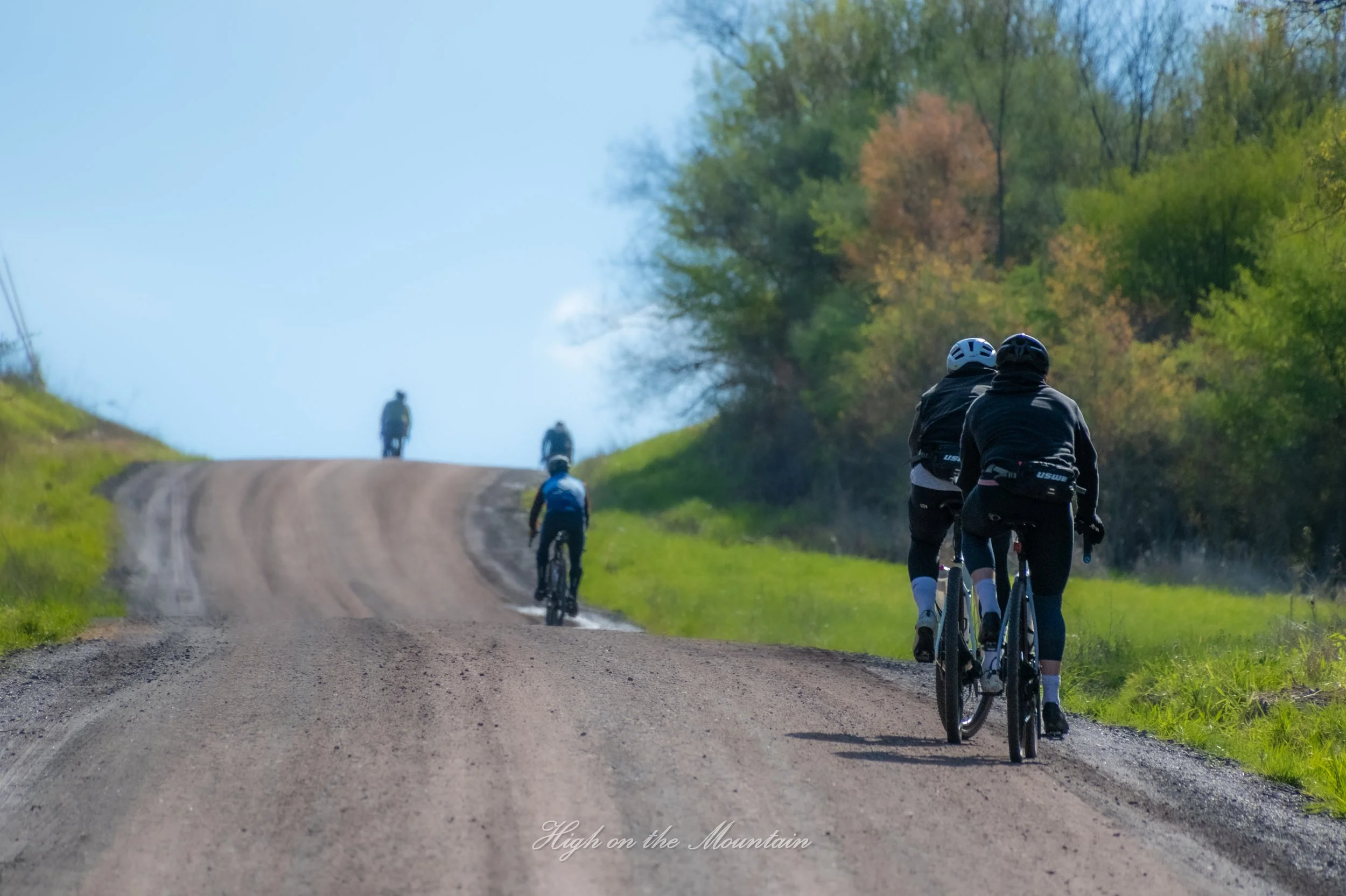 A group of cyclists riding on a dirt road through a green, rural landscape with trees and hills, with one cyclist ahead and others in the distance.