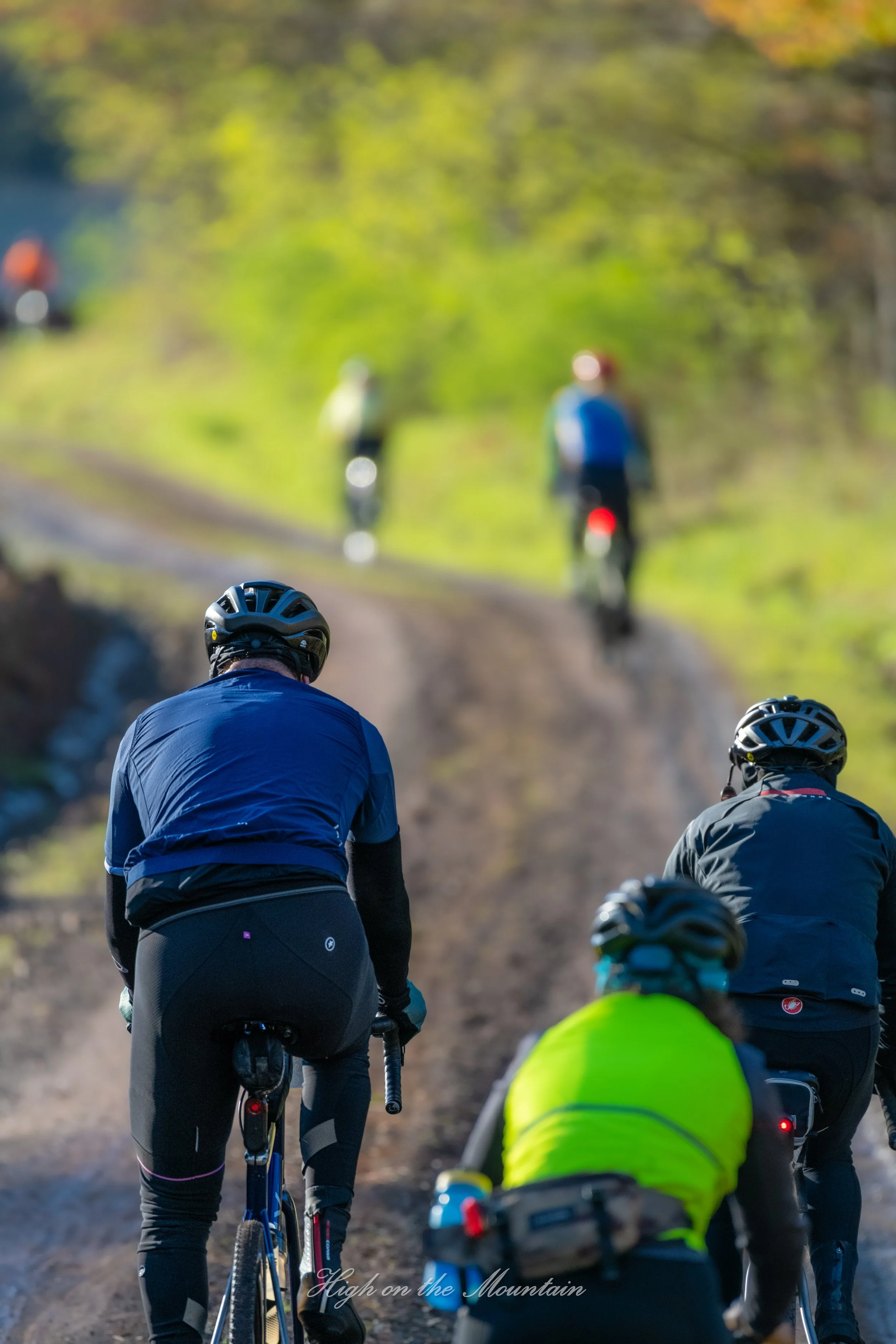 Group of cyclists riding mountain bikes on a dirt trail through a wooded area in daylight with green and orange leaves.