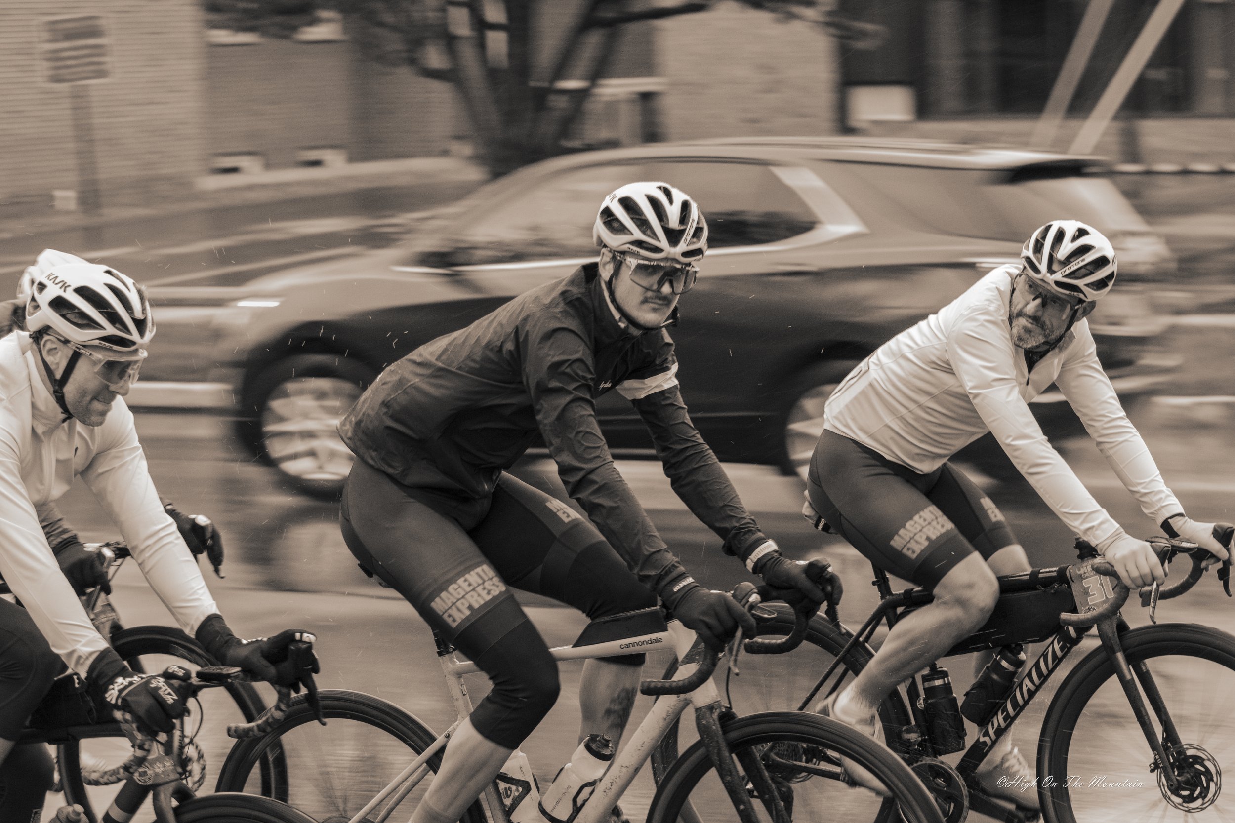 Three men wearing helmets and cycling gear riding bicycles on a city street with a blurred car in the background.