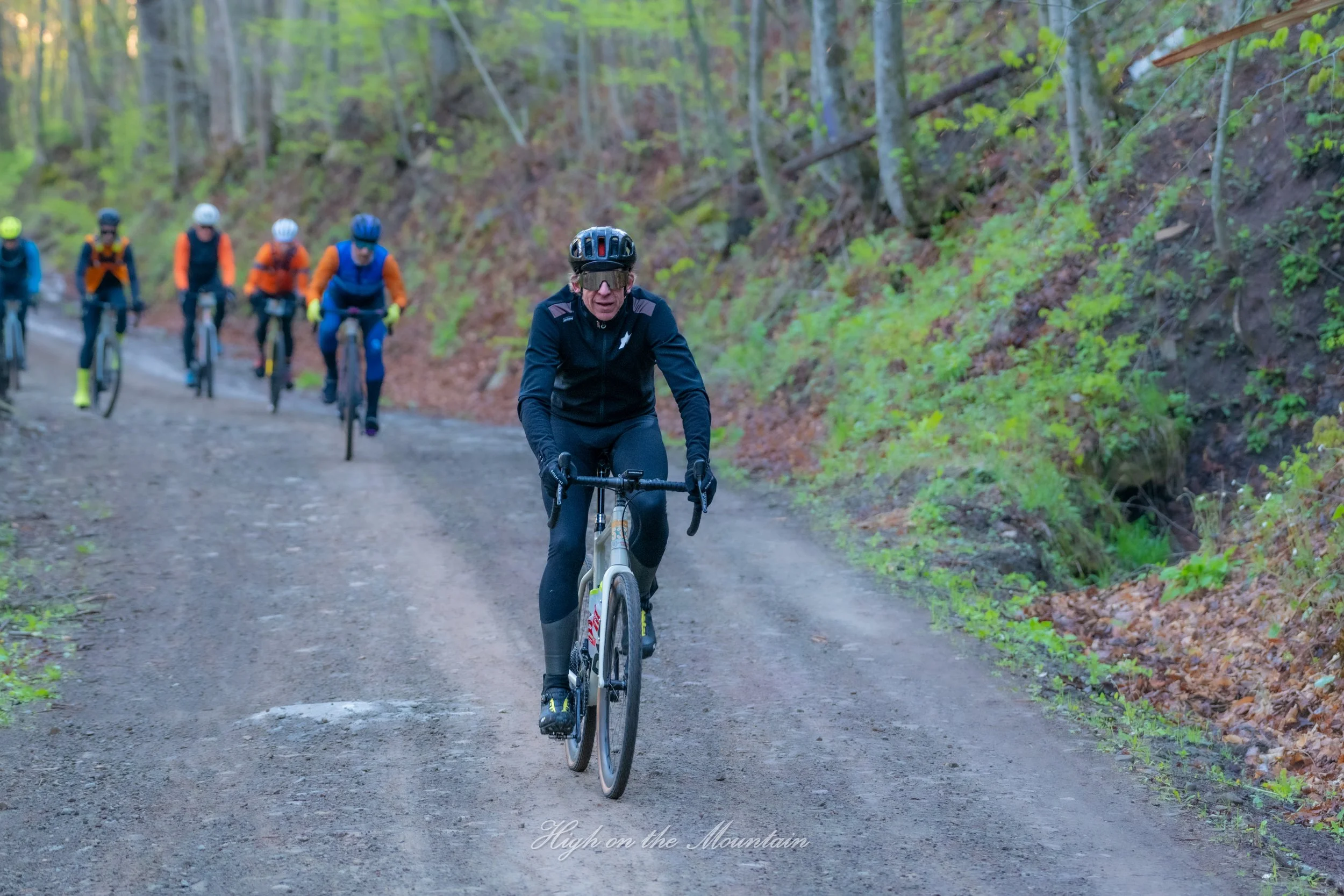 A group of cyclists riding on a dirt trail through a green forest, led by a woman in a black helmet and jacket, with the words "High on the Mountain" overlaid at the bottom.