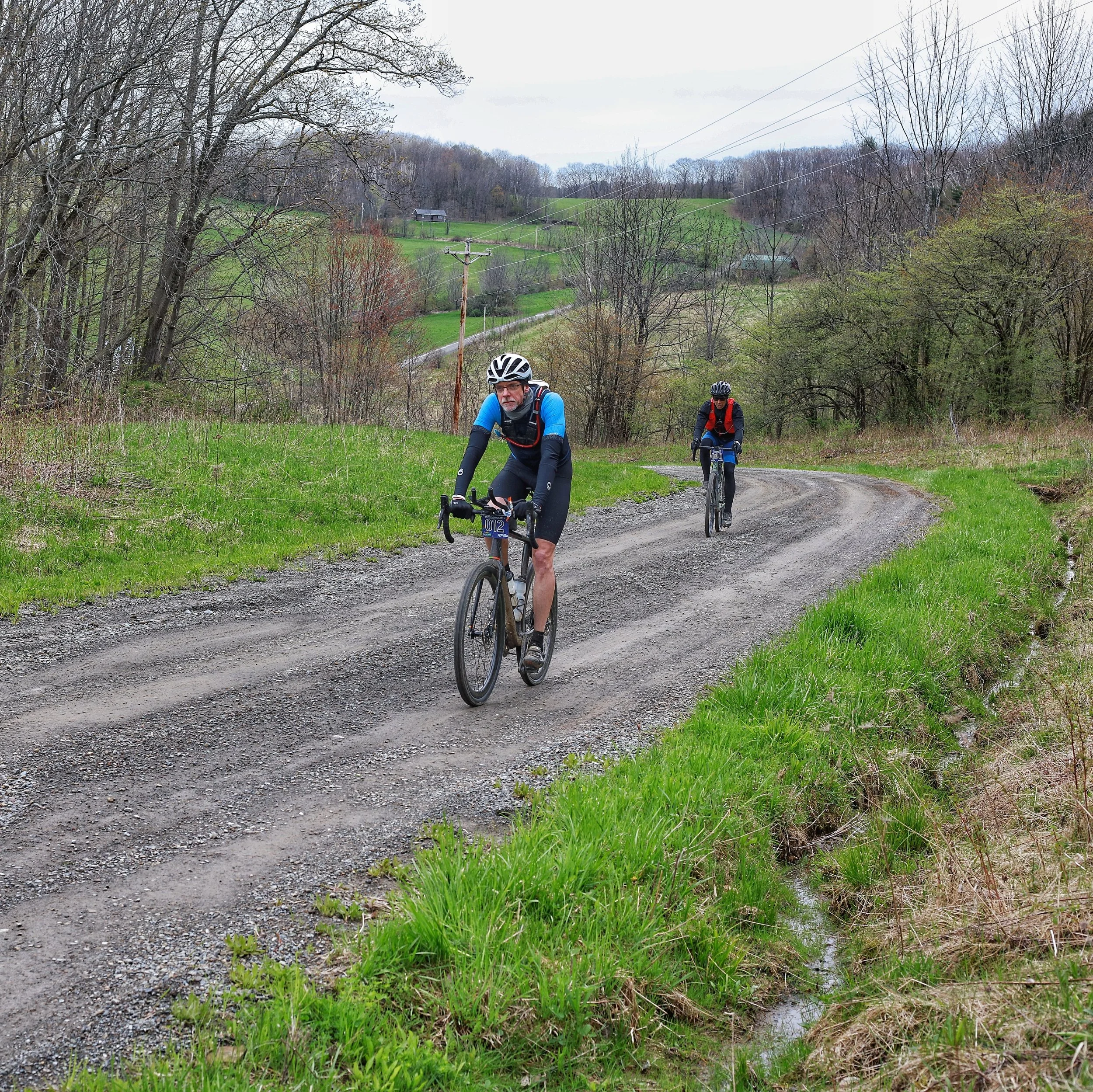 Two cyclists riding on a gravel country road surrounded by green grass and leafless trees, with rolling hills and power lines in the background.