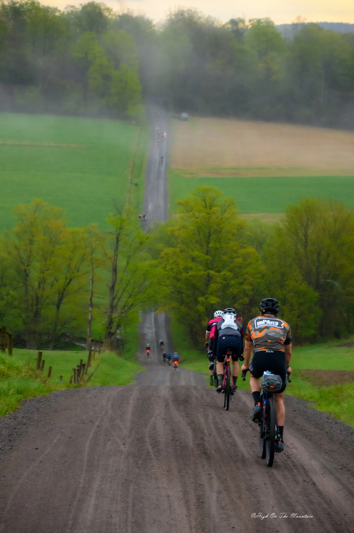 Cyclists biking on a rural dirt road through lush green countryside with trees, fields, and hills in the background during dusk.