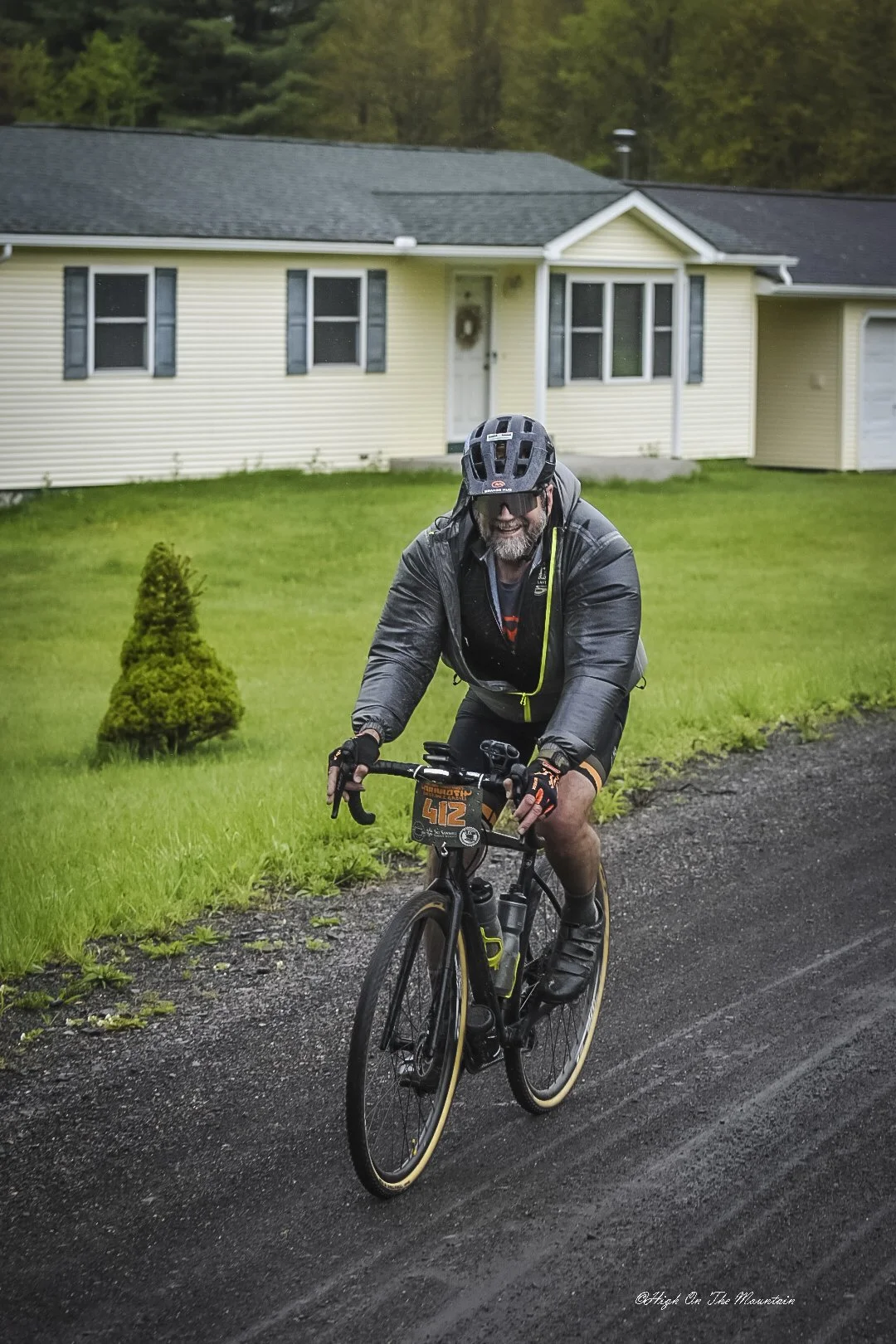 A man riding a mountain bike on a dirt path in a residential area, wearing a black helmet, sunglasses, a black jacket, and gloves, smiling and looking forward.