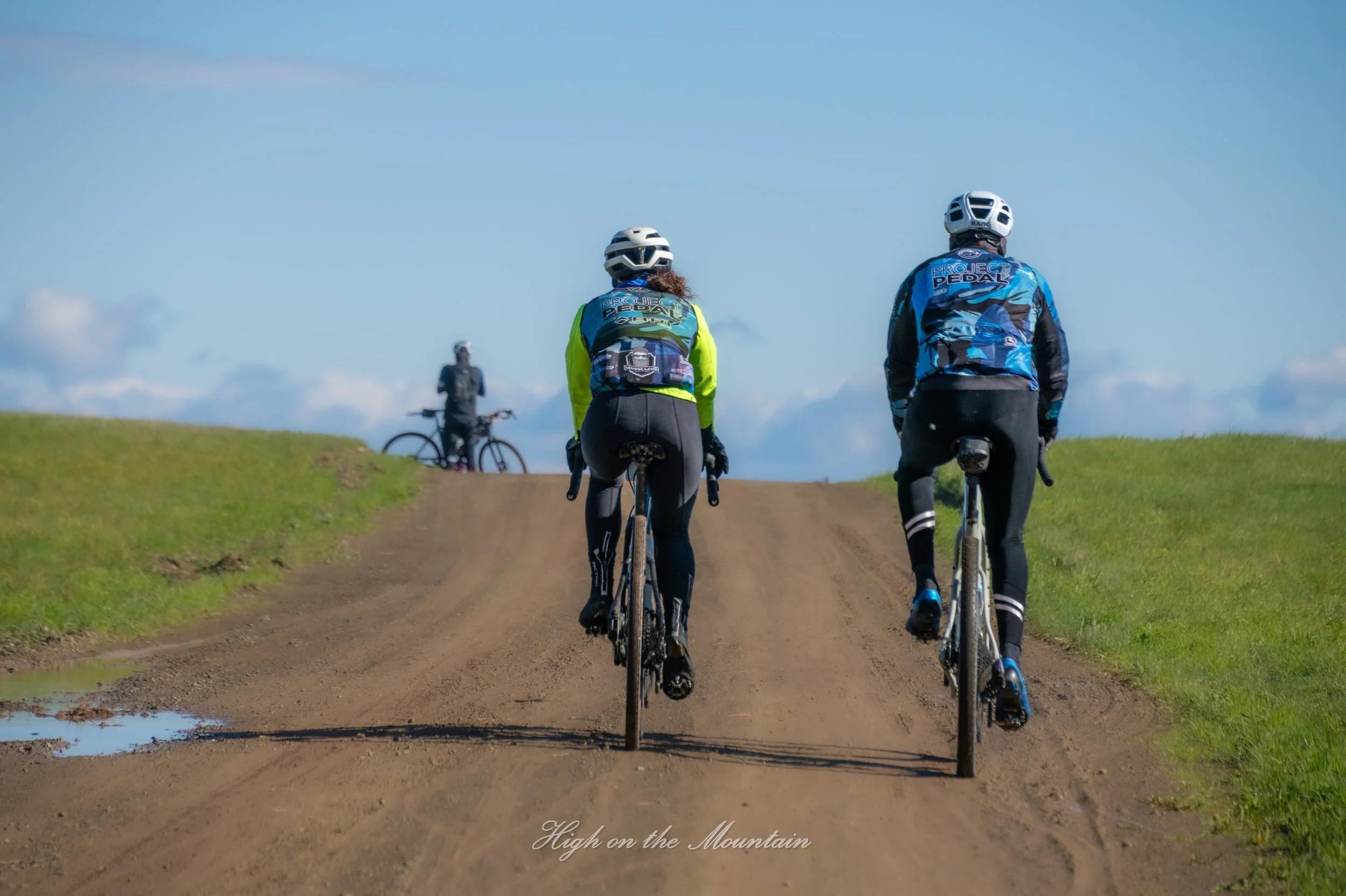 Two cyclists wearing helmets and colorful jerseys riding mountain bikes on a dirt trail, with another cyclist visible in the background, under a blue sky with some clouds, on a grassy hillside.