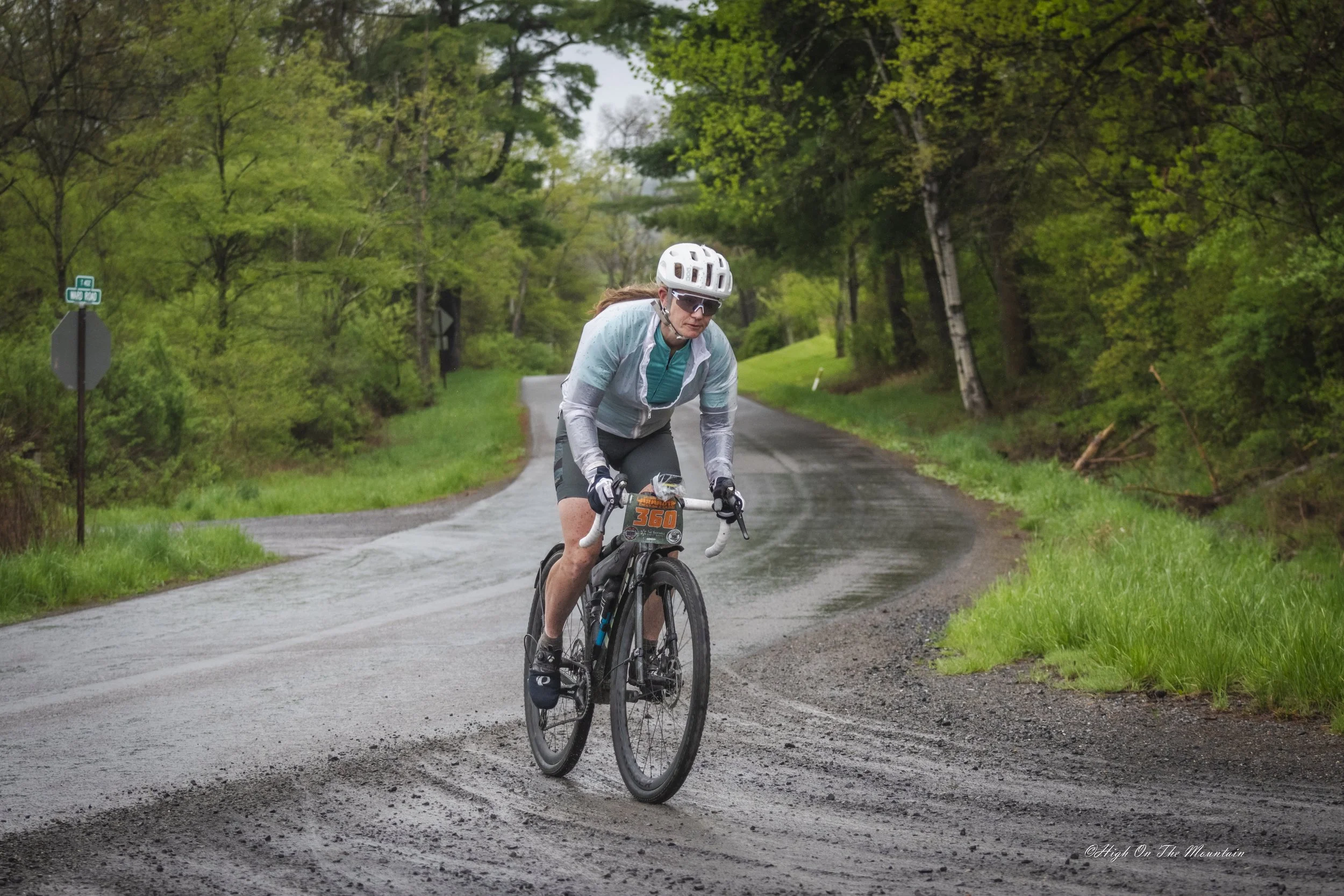A person riding a mountain bike on a rural, wet road surrounded by green trees during rainy weather.