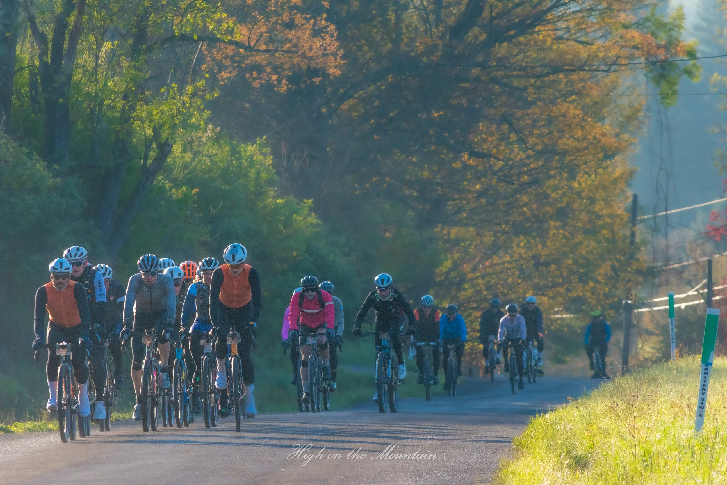 A group of cyclists riding on a paved path through a forested area with green and orange autumn foliage, with three volunteers walking beside them, against a backdrop of trees and a partly cloudy sky.