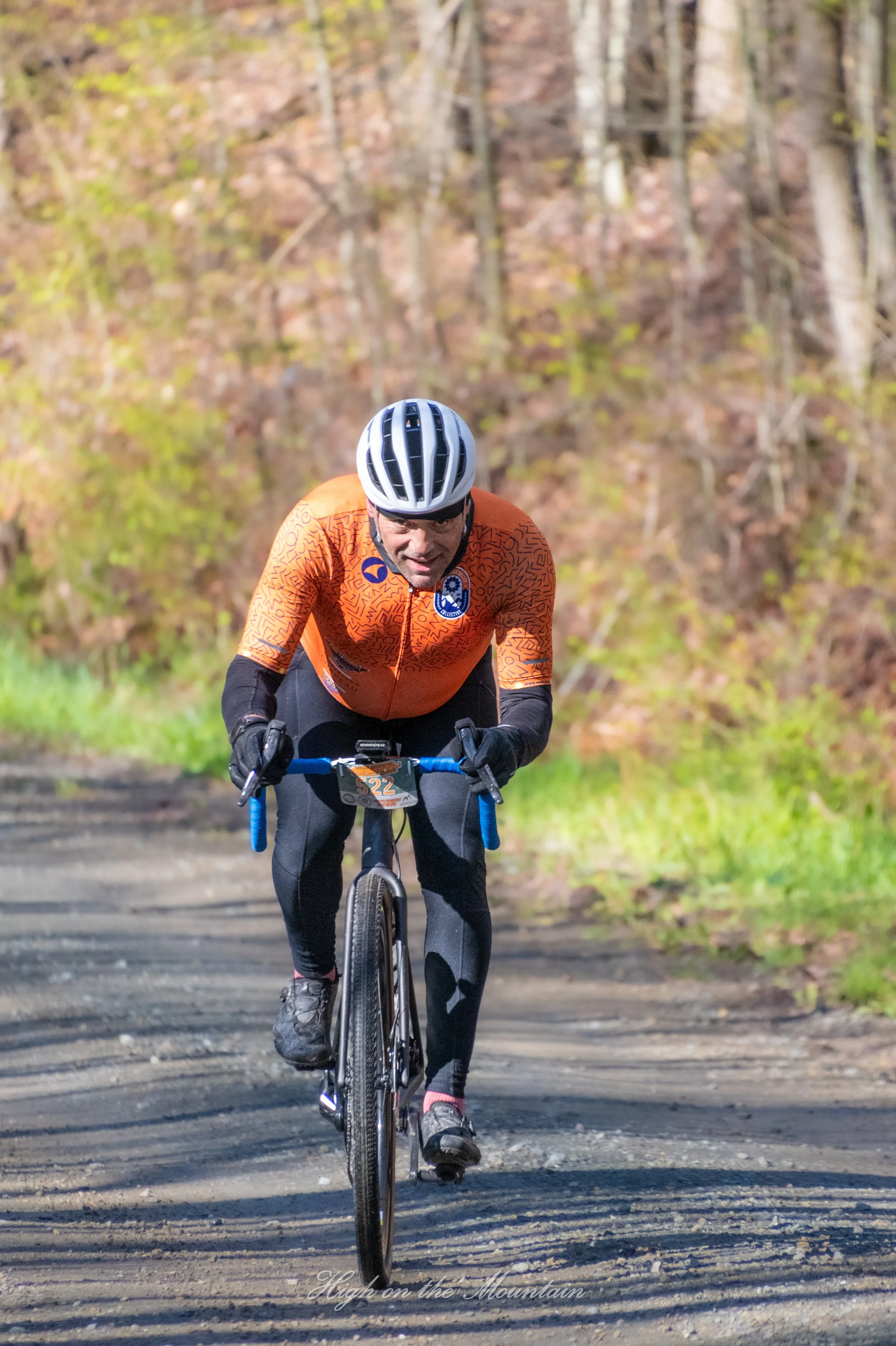 A man wearing a helmet and orange cycling jersey riding a mountain bike on a dirt trail in a forest with trees and autumn leaves.
