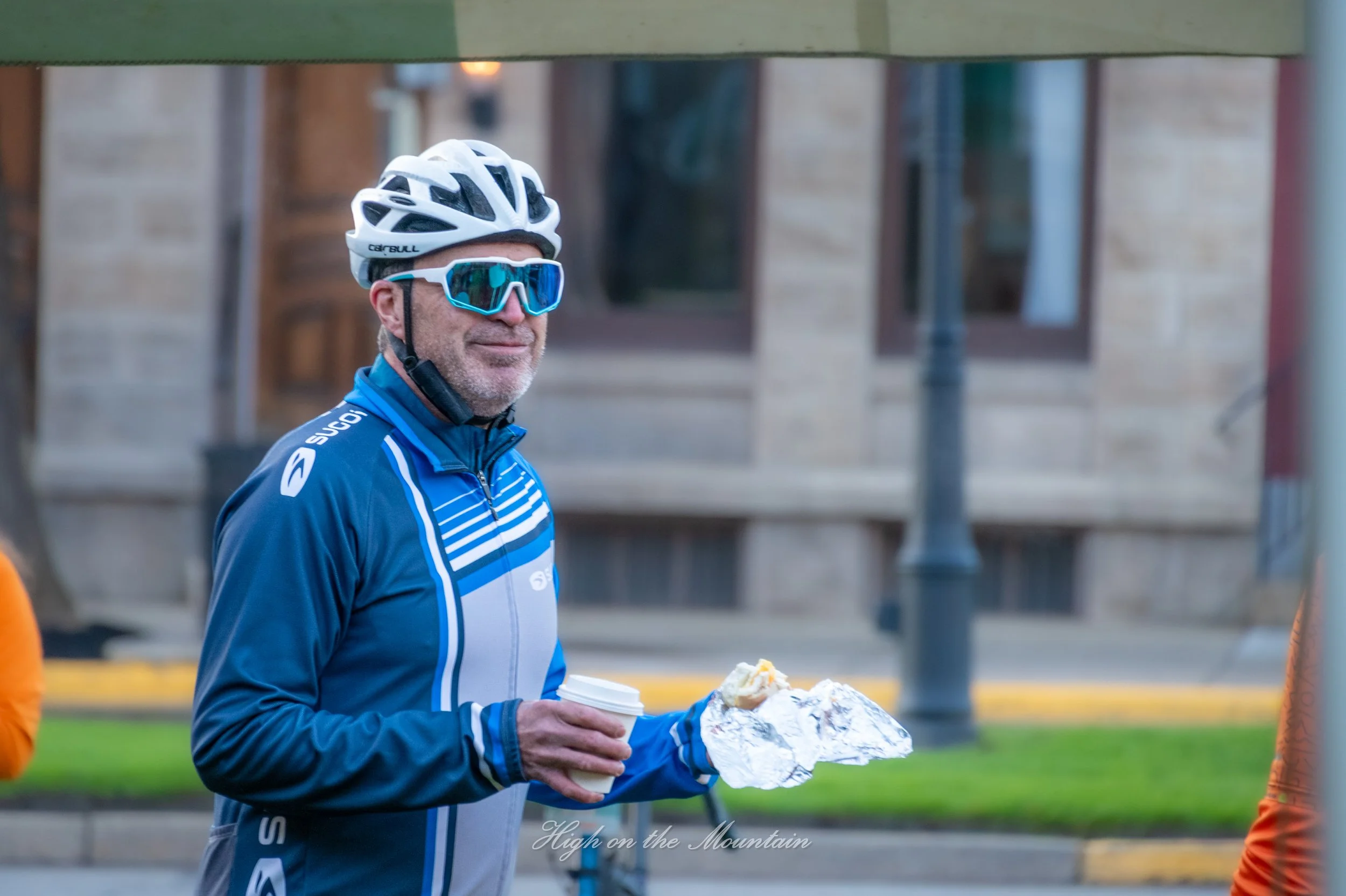 A man in a cycling helmet, sunglasses, and blue cycling jacket holding a coffee cup and a sandwich in an outdoor urban setting.