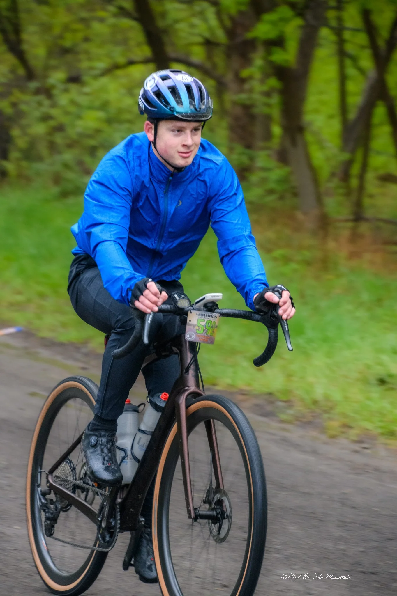 A person wearing a blue jacket and a helmet riding a mountain bike on a forest trail. Trees and greenery in the background.