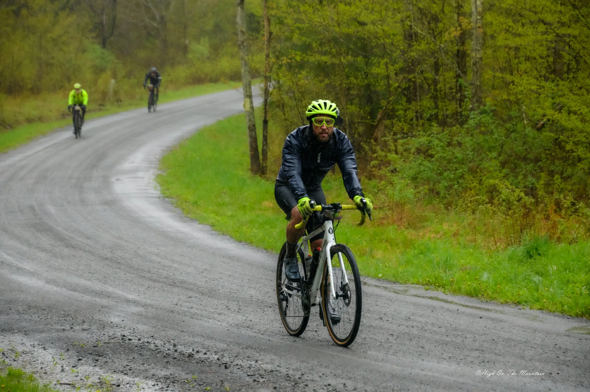 A man wearing a bright yellow helmet, glasses, and a black rain jacket rides a white bike on a wet, winding gravel road surrounded by green trees in a forested area. Two other cyclists are visible in the background.