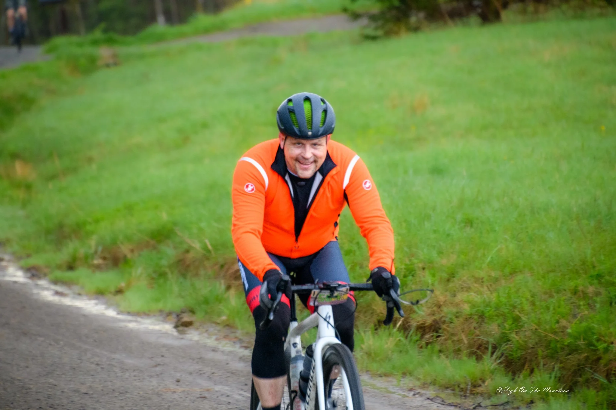 A man wearing an orange jacket and a black helmet riding a mountain bike on a dirt trail through green grass and trees.