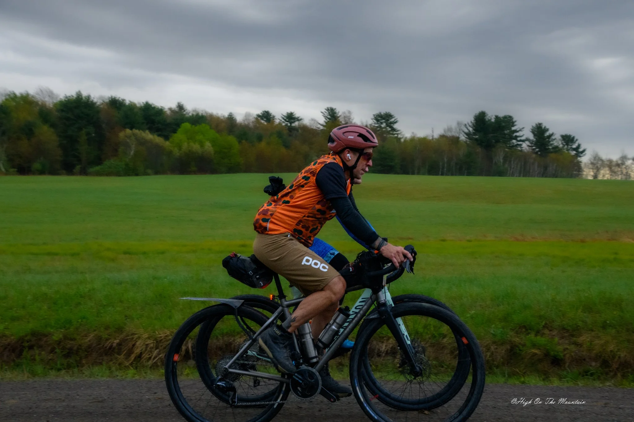 A person riding a bicycle on a trail through a grassy field with a background of trees and an overcast sky.