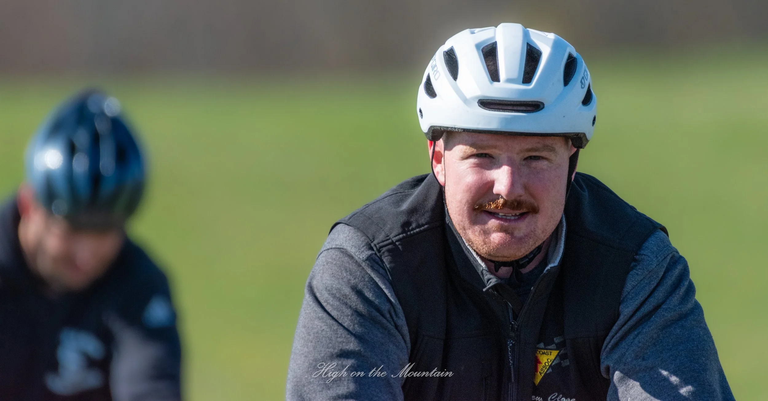 A man wearing a white bicycle helmet and a dark jacket, outdoors on a grassy field, smiling and looking to the side. There is another person blurred in the foreground also wearing a helmet.