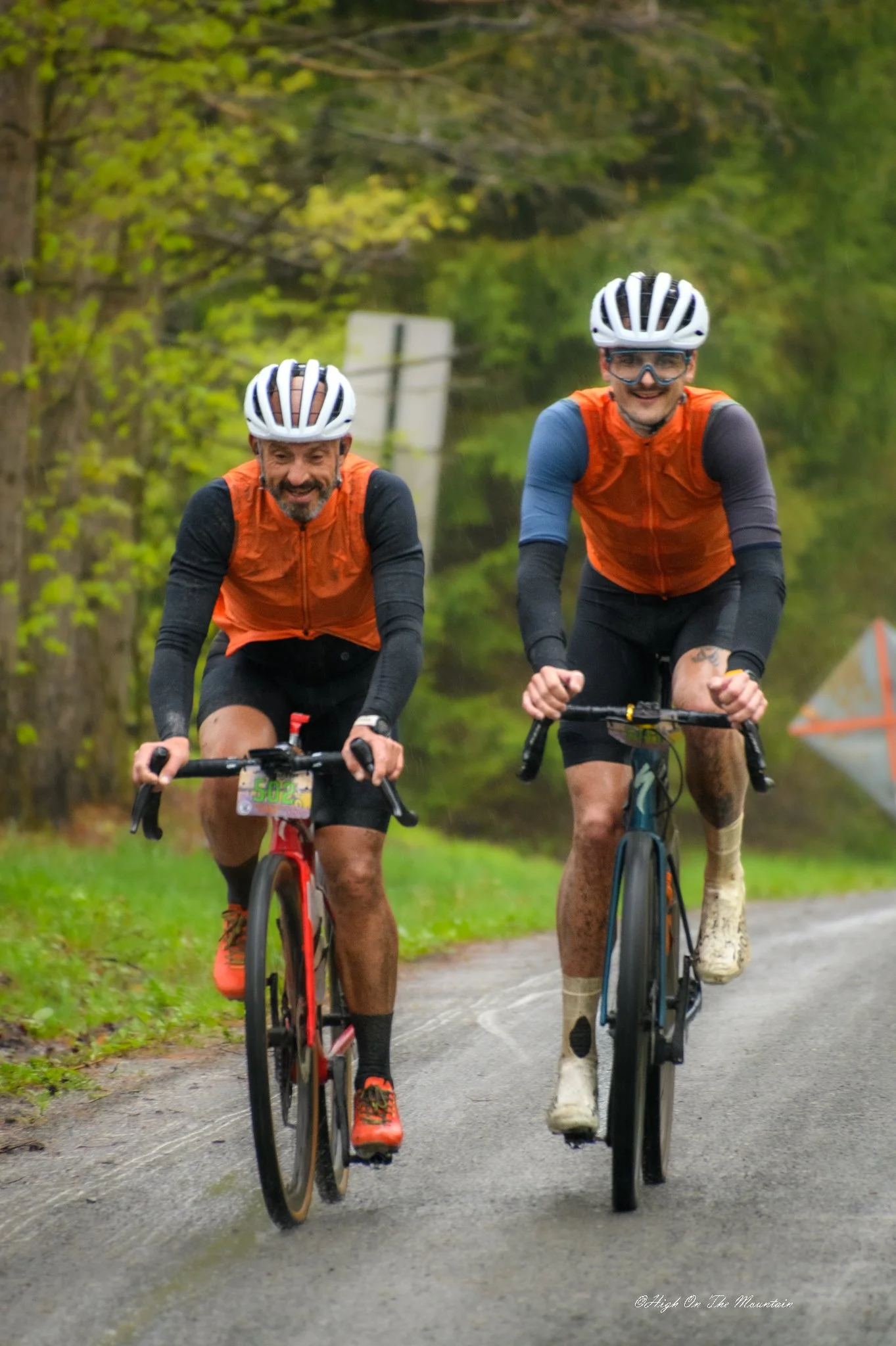 Two men riding mountain bikes on a trail in a wooded area, wearing helmets and orange vests, smiling and enjoying the ride.