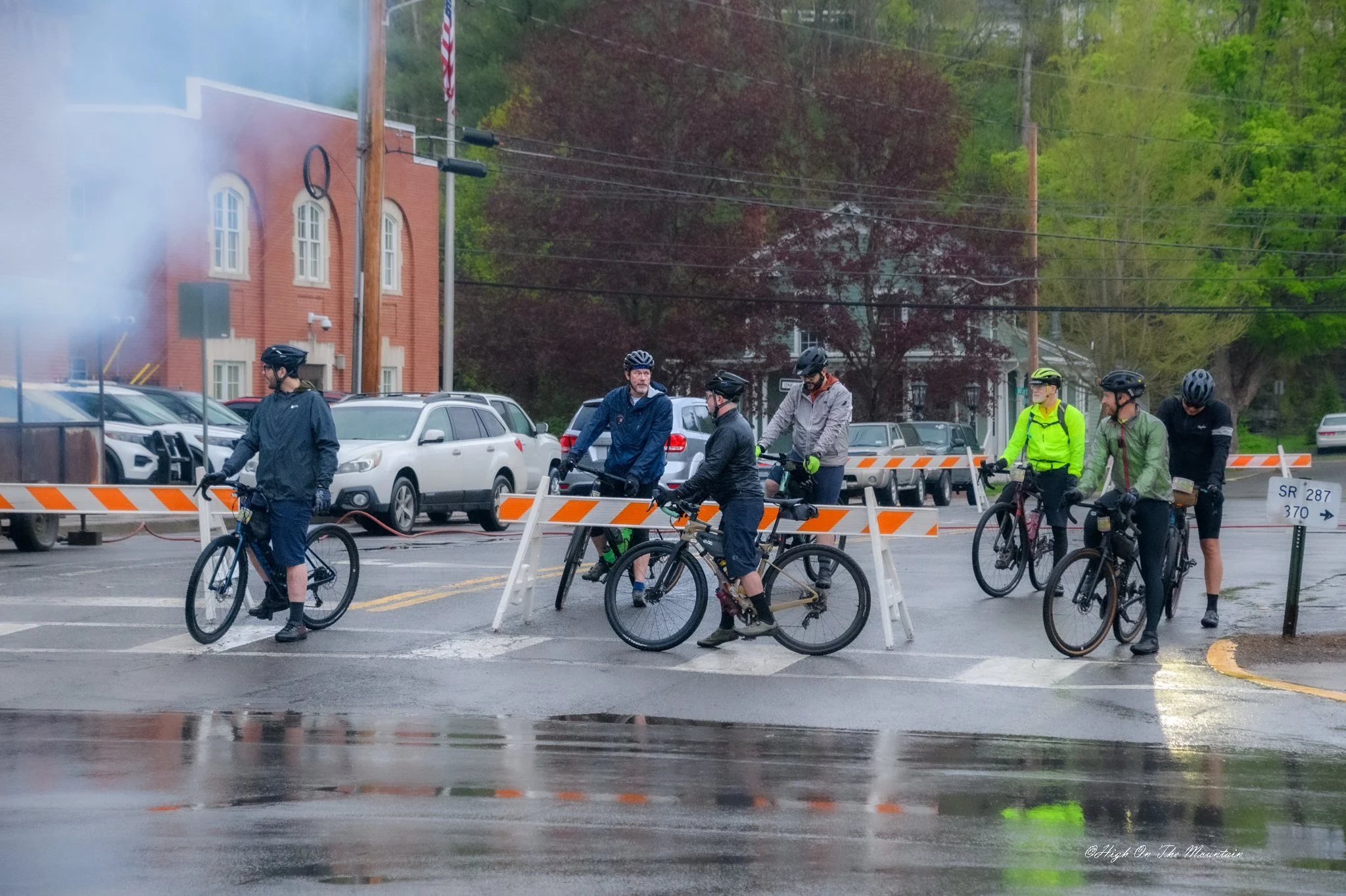 A group of cyclists stopped on a wet street behind a construction barrier, with parked cars, trees, and buildings in the background.