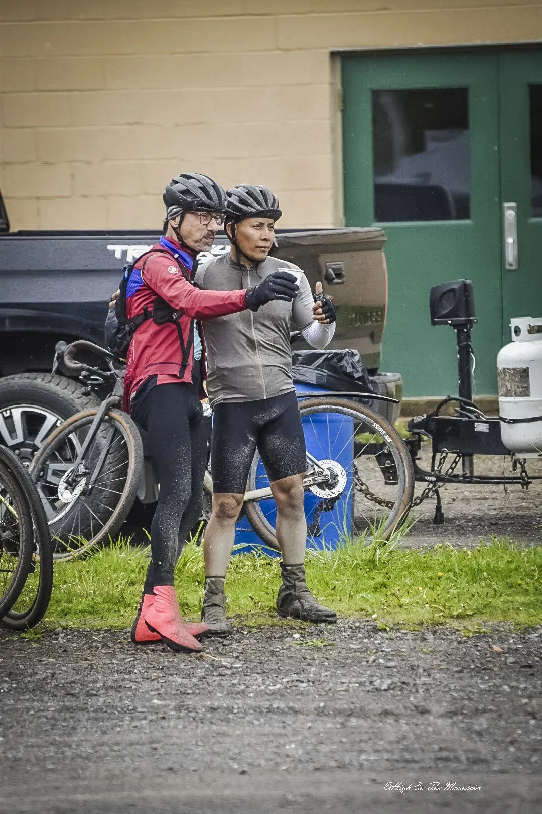 Two cyclists in cycling gear and helmets standing outdoors, looking at a smartphone together. They are dirty from riding, with wet and muddy legs, and are near bikes and support vehicles.