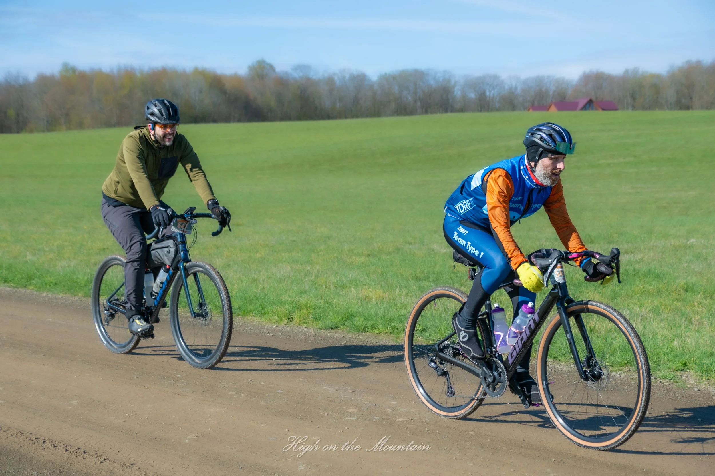 Two middle-aged men riding bicycles outdoors on a dirt path in a rural area with green fields and trees in the background under a partly cloudy sky.