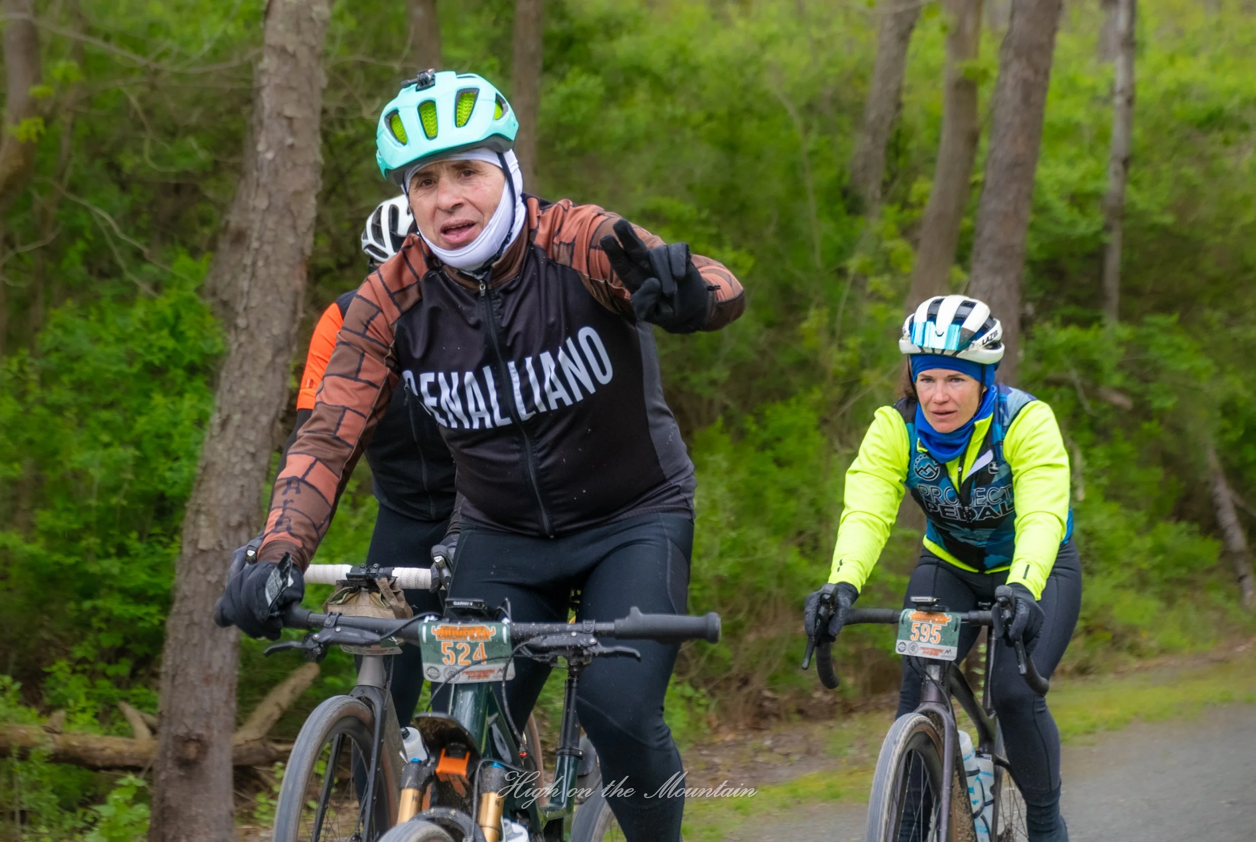 Two mountain bikers riding through a wooded trail, with one man making a peace sign and the woman riding behind him, both wearing helmets, gloves, and cycling gear.