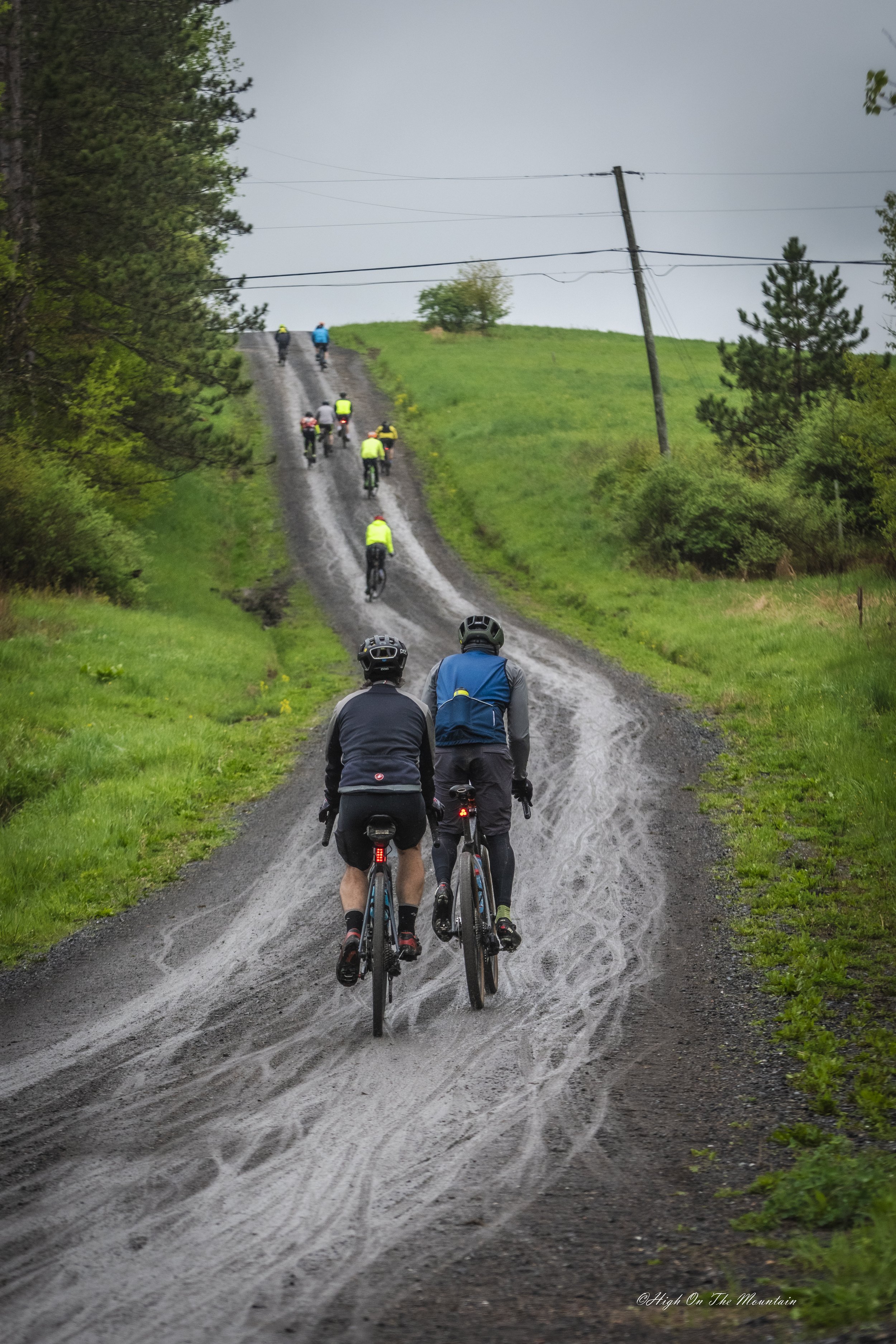 Two cyclists riding on a gravel dirt road with a group of cyclists further ahead, surrounded by green grass and trees on a cloudy day.