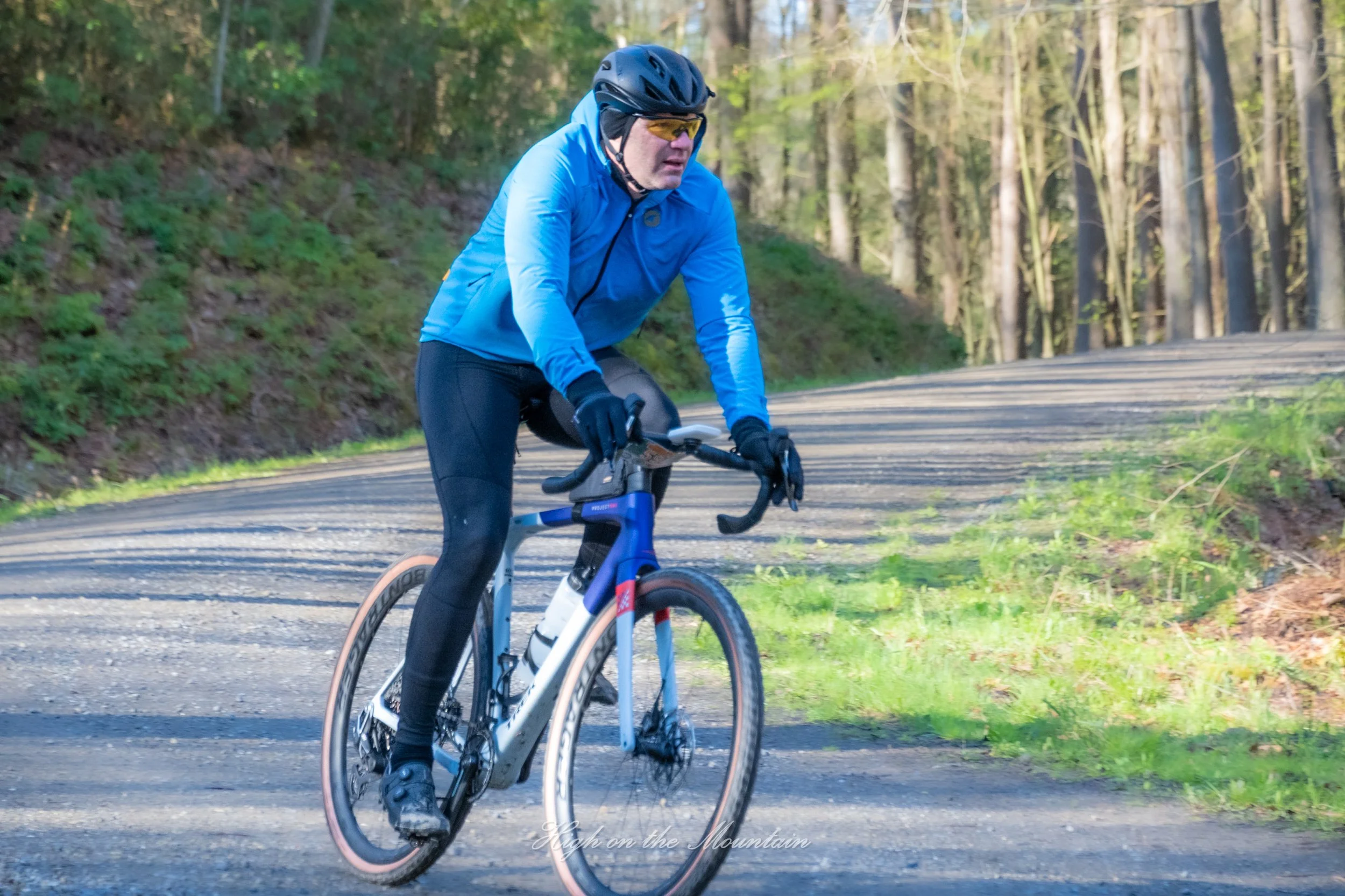 A man in a blue jacket and black helmet riding a bicycle on a forested trail with trees and greenery in the background.