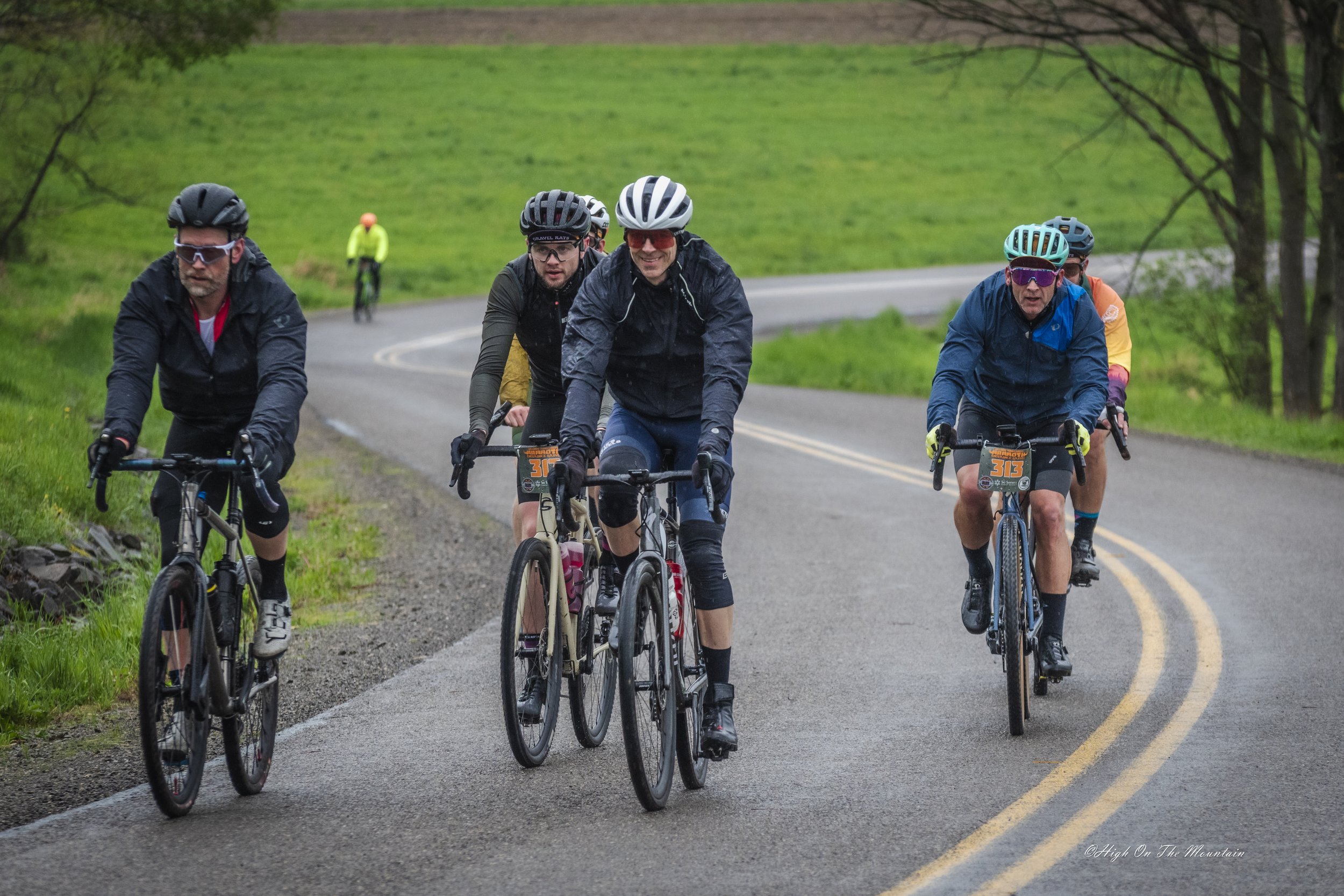 Group of five cyclists riding on a winding rural road with green grass and trees around, and a cyclist in bright clothing in the background.