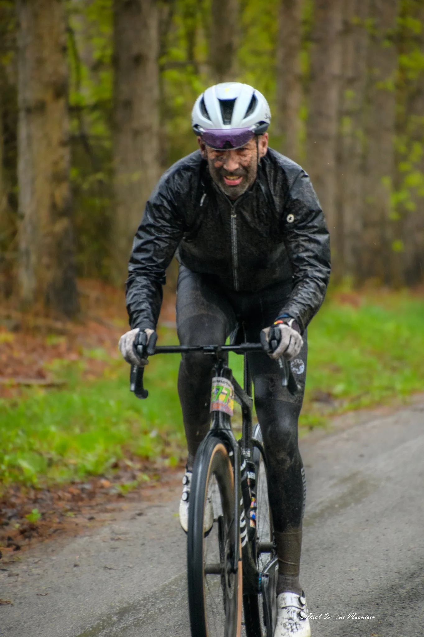 A man wearing a helmet and black rain jacket riding a bicycle on a wet trail in a forest with green trees.