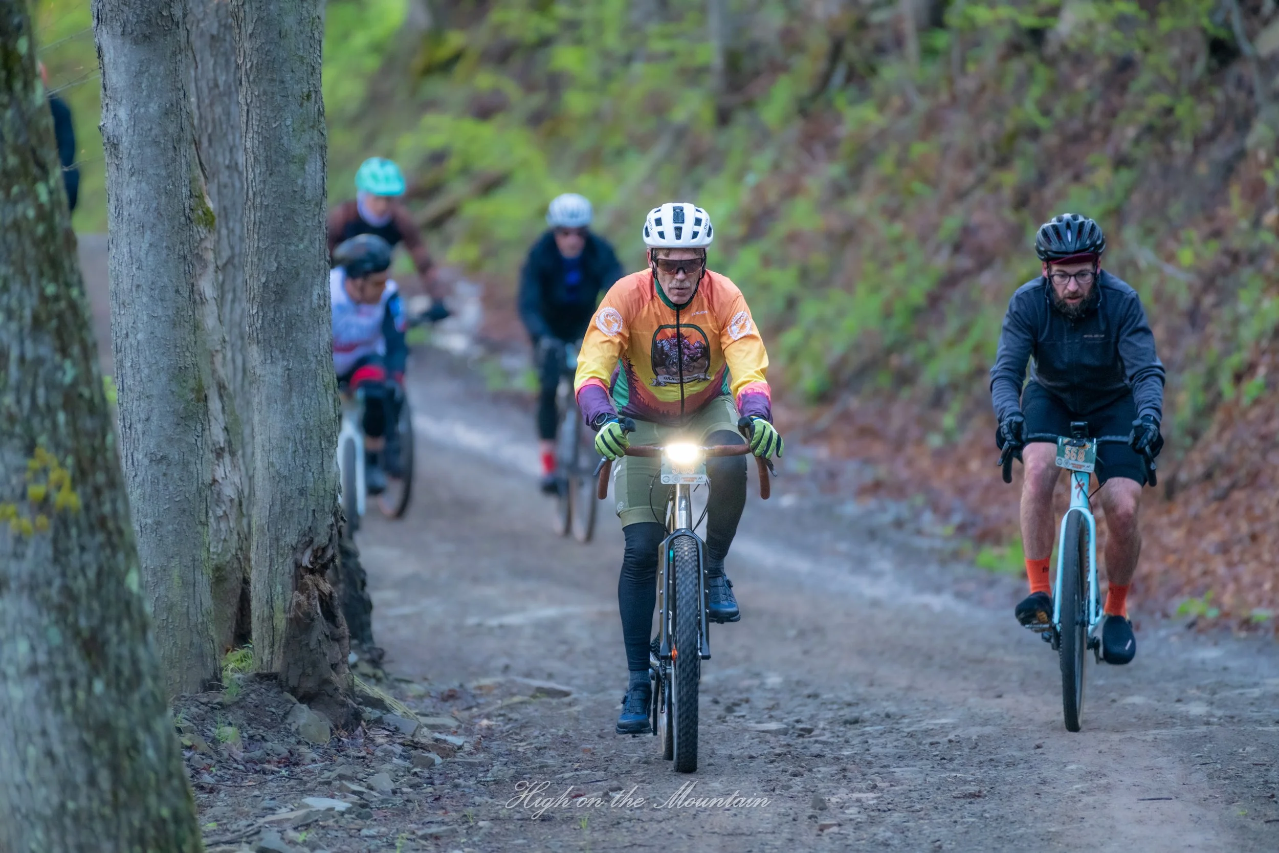 Group of mountain bikers riding on a forest trail surrounded by trees and greenery, with a focus on an older man wearing a white helmet and sunglasses leading the ride.