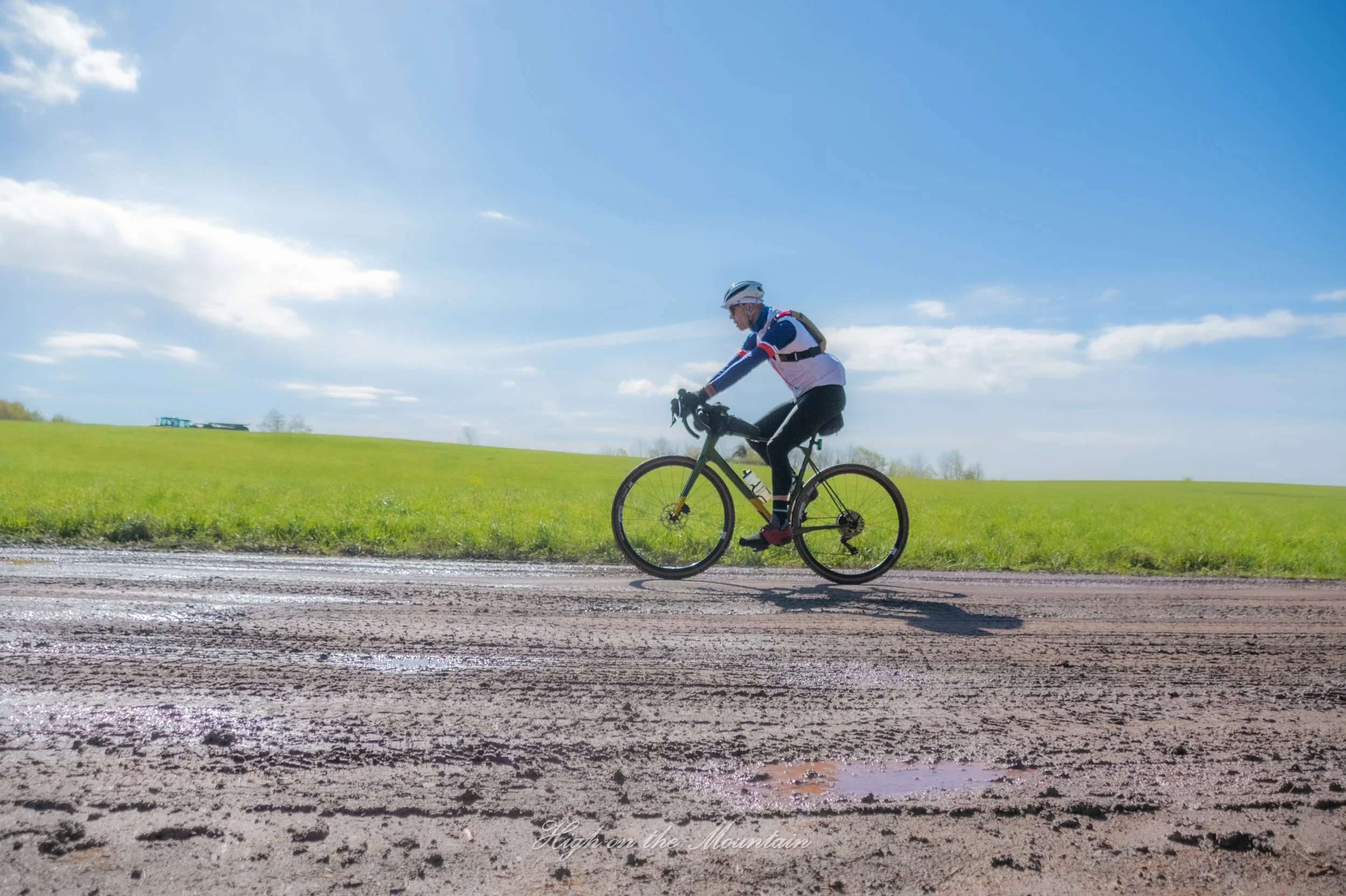 A person riding a bicycle on a dirt road through a grassy landscape with a blue sky and some clouds.