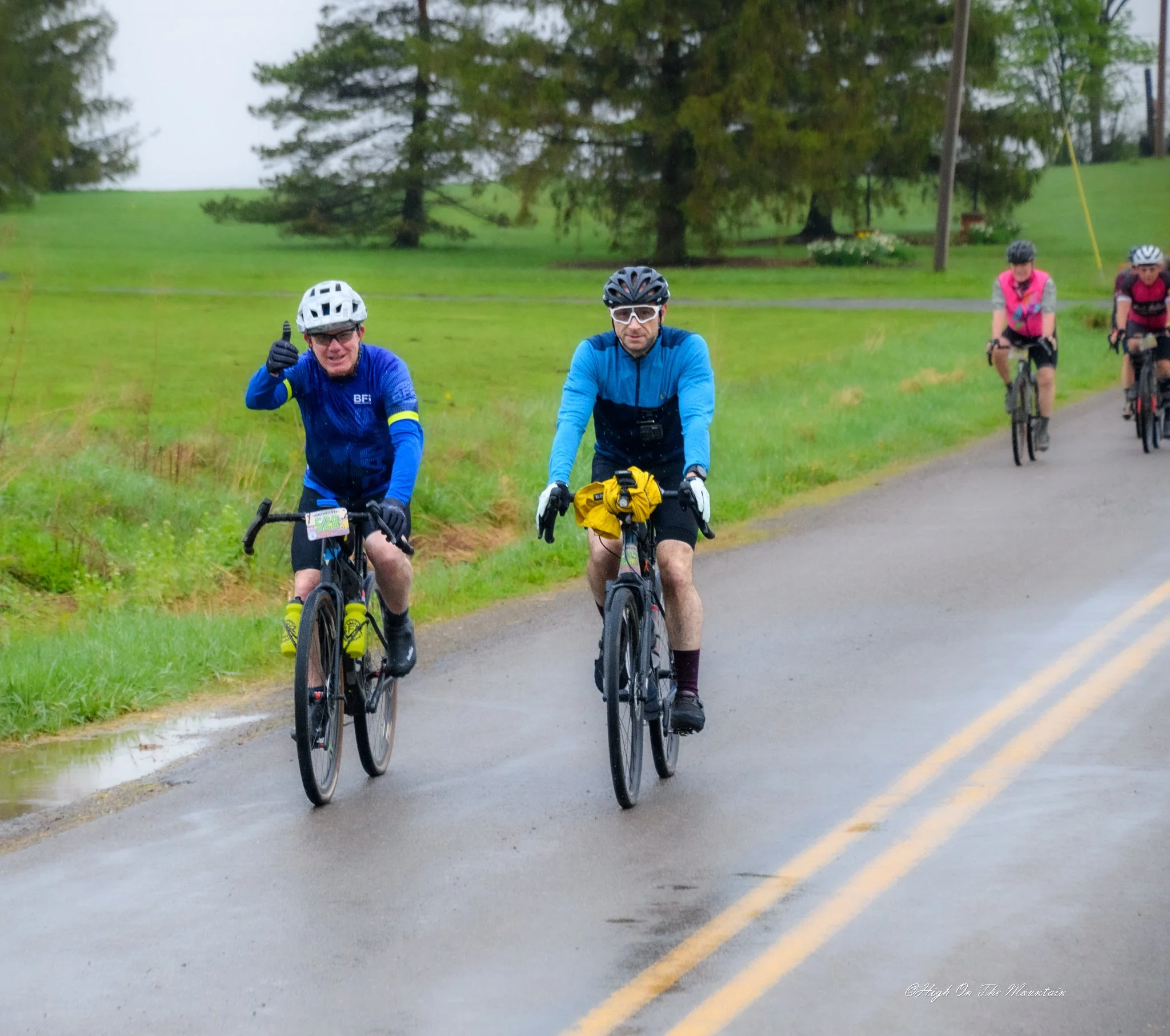 Three cyclists riding on a rainy day on a rural road with greenery and trees around.