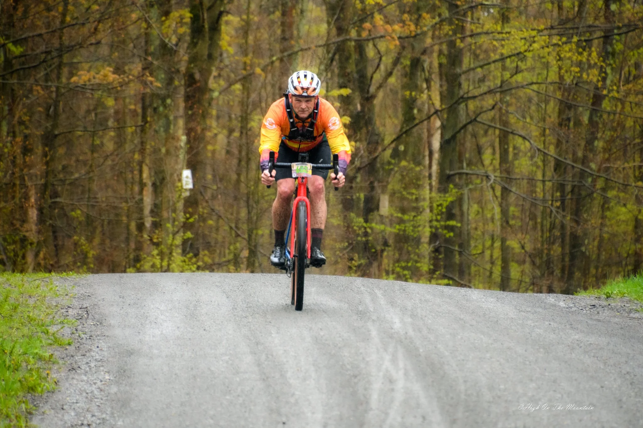A man riding a mountain bike on a gravel trail through a forest with trees showing fall foliage.