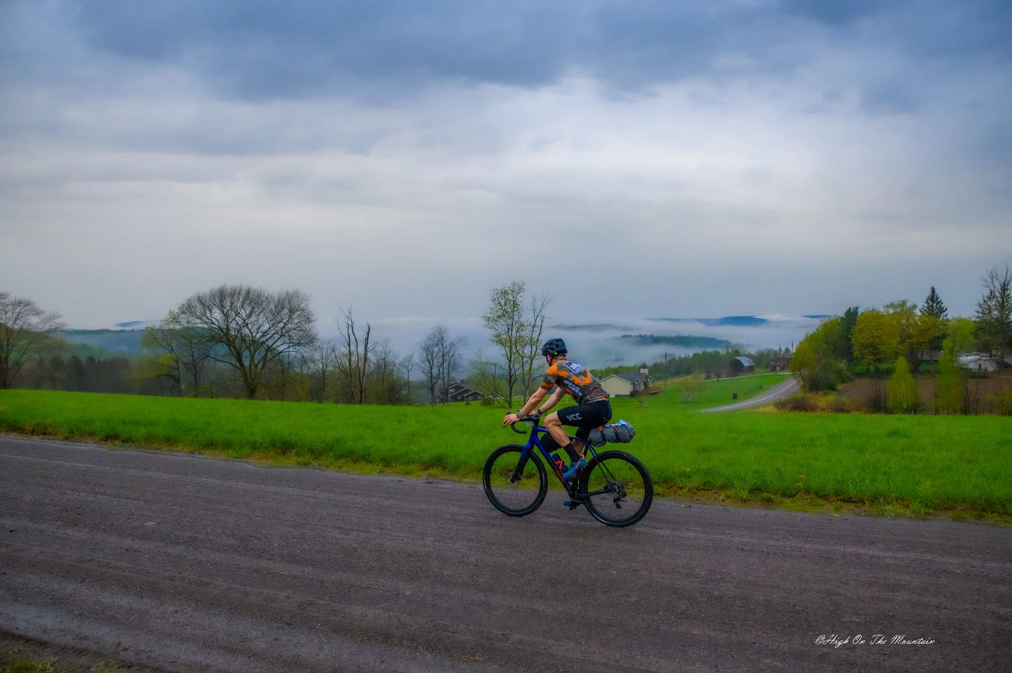 A cyclist wearing a helmet and cycling gear rides along a road in a rural landscape with green grass, trees, and houses, under a cloudy sky with mist in the background.