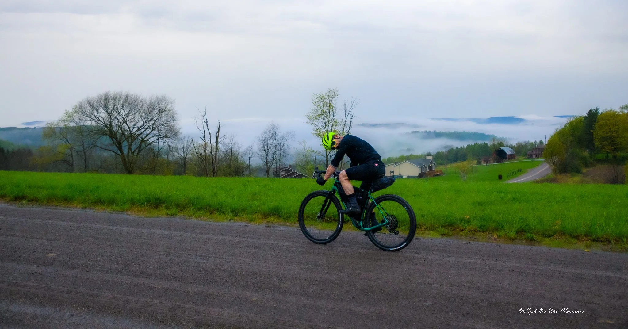 A cyclist in black riding a mountain bike on a dirt road in a rural landscape with green grass, trees, and houses in the background, under a cloudy sky.